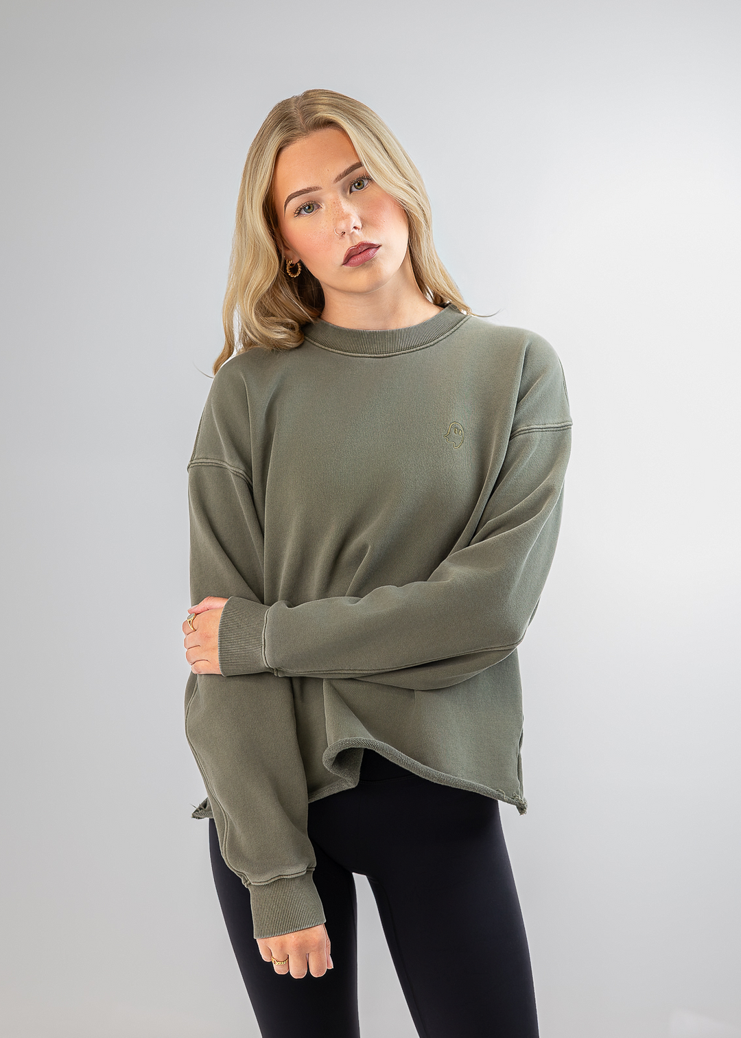 A woman modeling an oversized crewneck sweatshirt in olive green, paired with black leggings, against a light gray studio background.