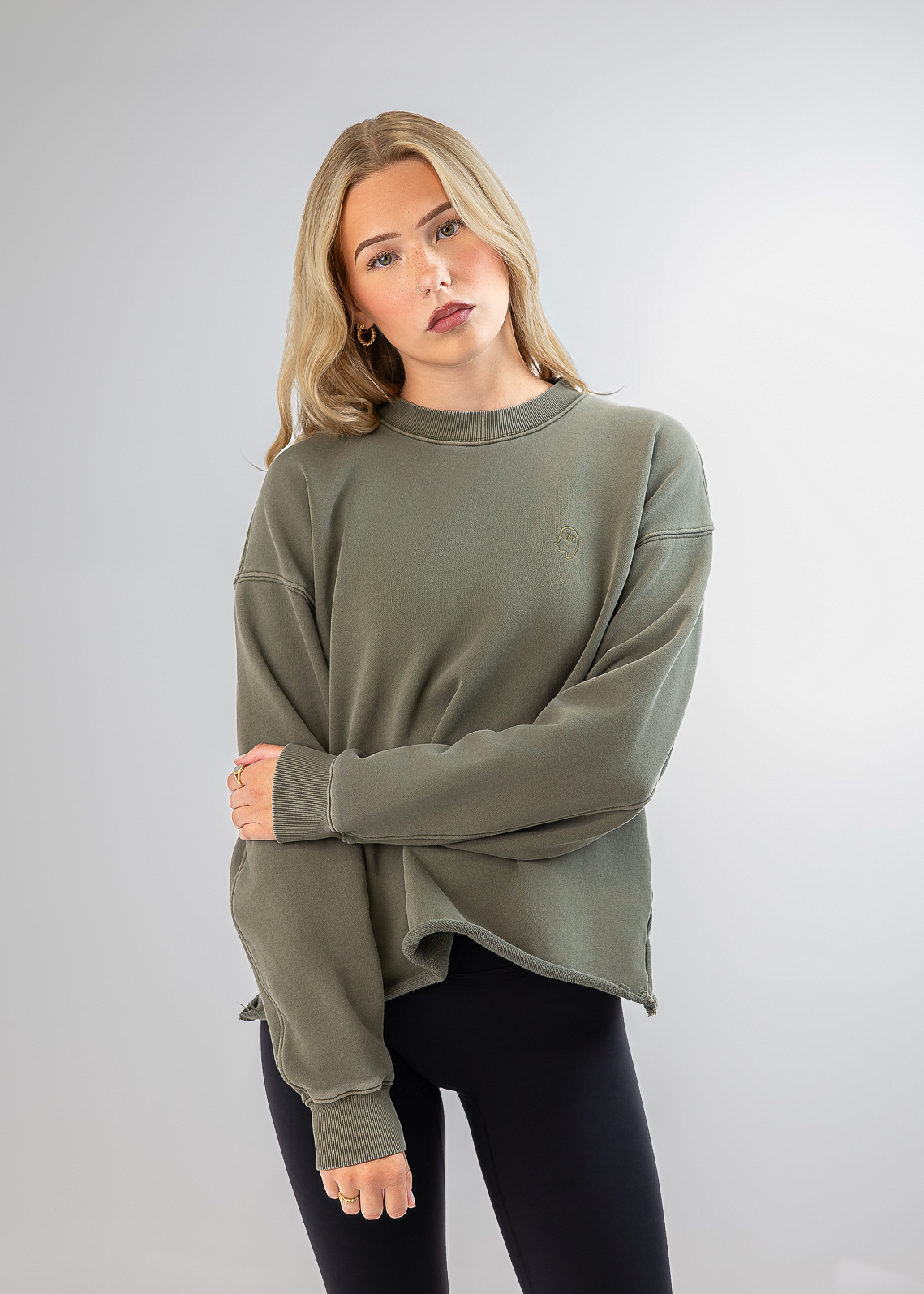 A woman modeling an oversized crewneck sweatshirt in olive green, paired with black leggings, against a light gray studio background.