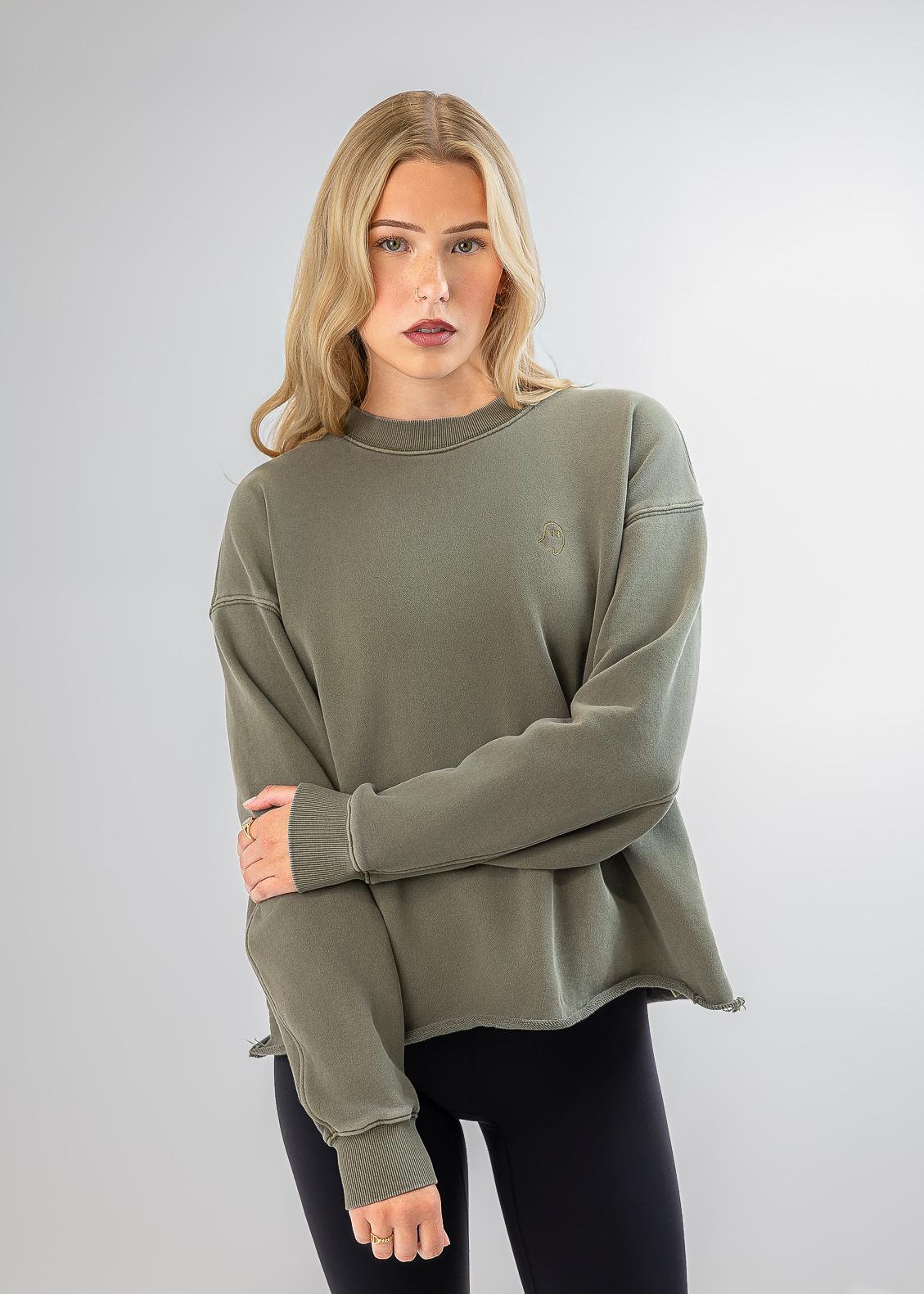 A blonde model wearing an oversized sage green crewneck sweatshirt and black leggings, standing against a light gray studio background.