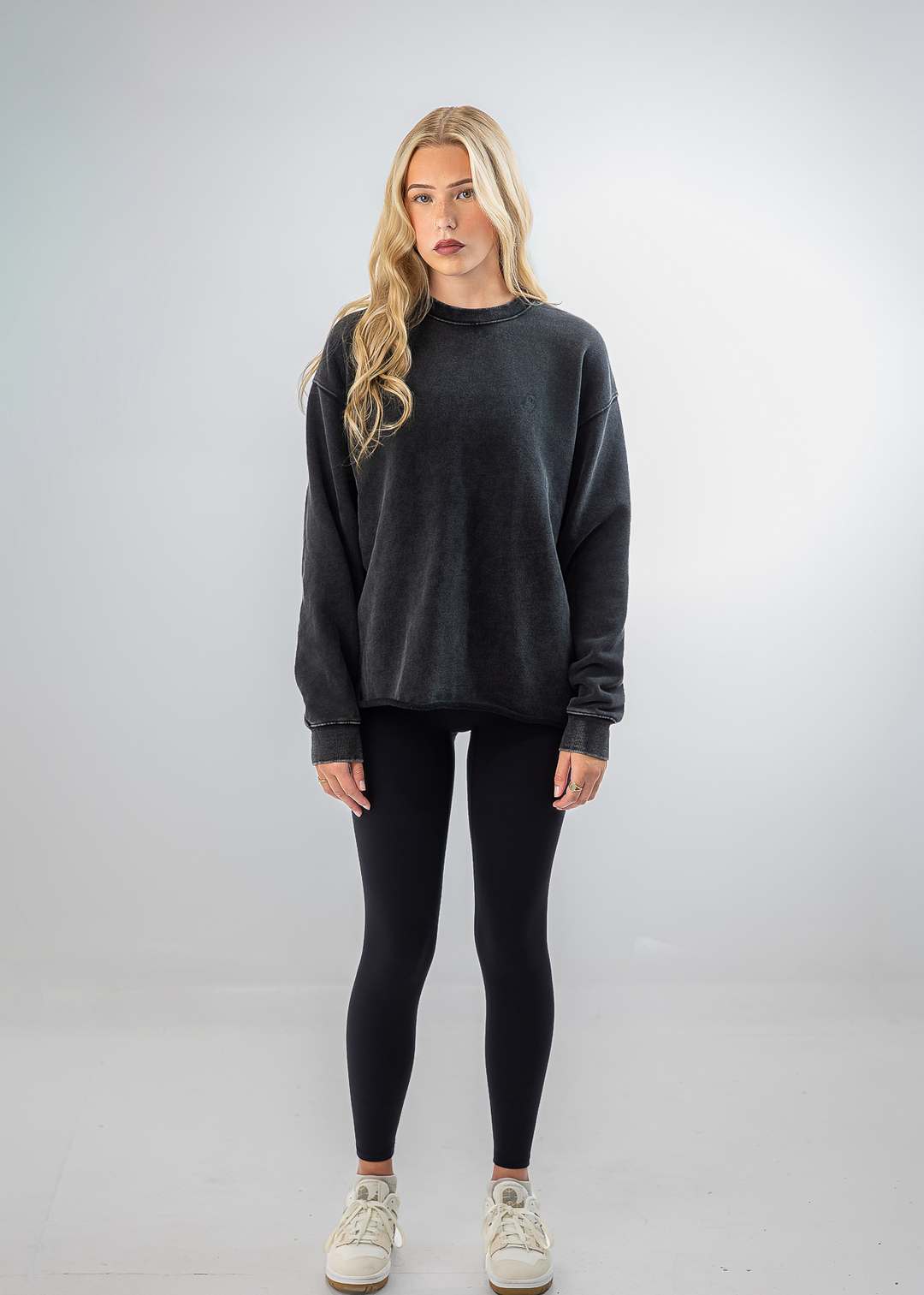 A woman stands against a light gray studio background wearing an oversized charcoal gray crewneck sweatshirt, black leggings, and off-white sneakers.