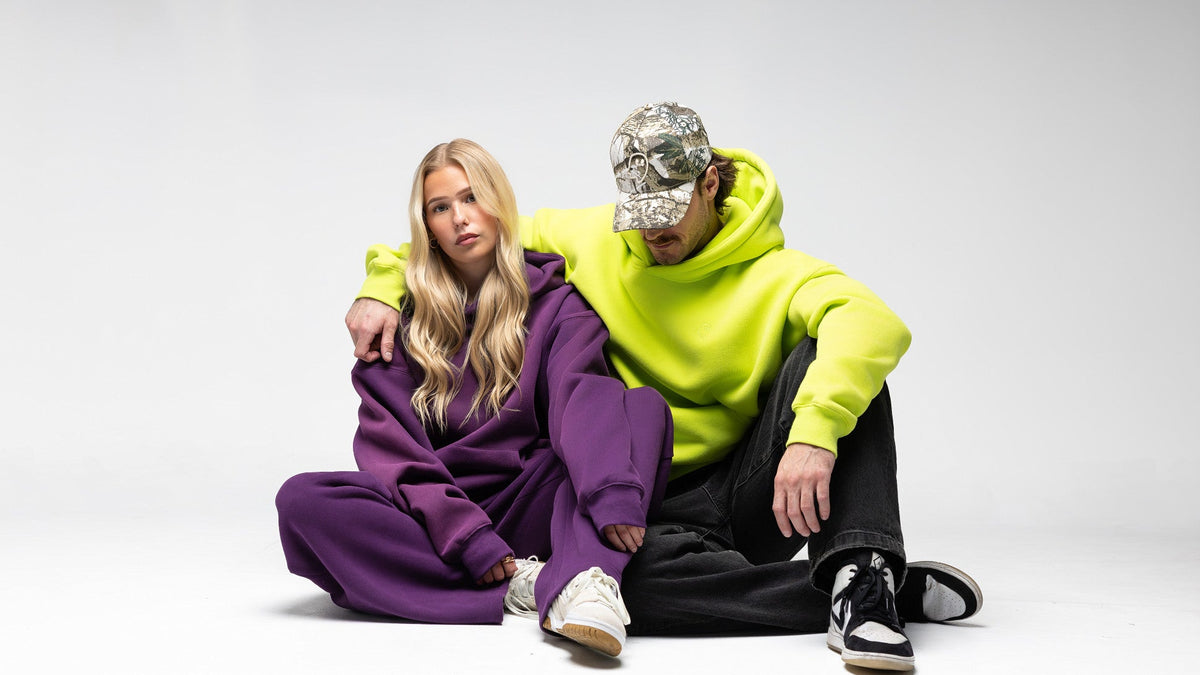 A man and a woman sit together on a light gray studio floor. The woman wears an oversized purple sweatshirt and matching pants with white sneakers.