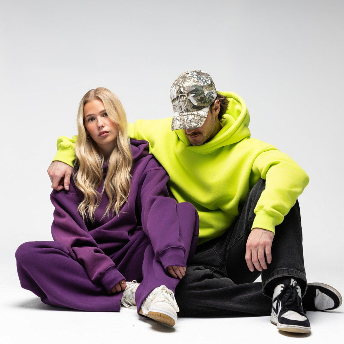 A man and a woman sit together on a light gray studio floor. The woman wears an oversized purple sweatshirt and matching pants with white sneakers.