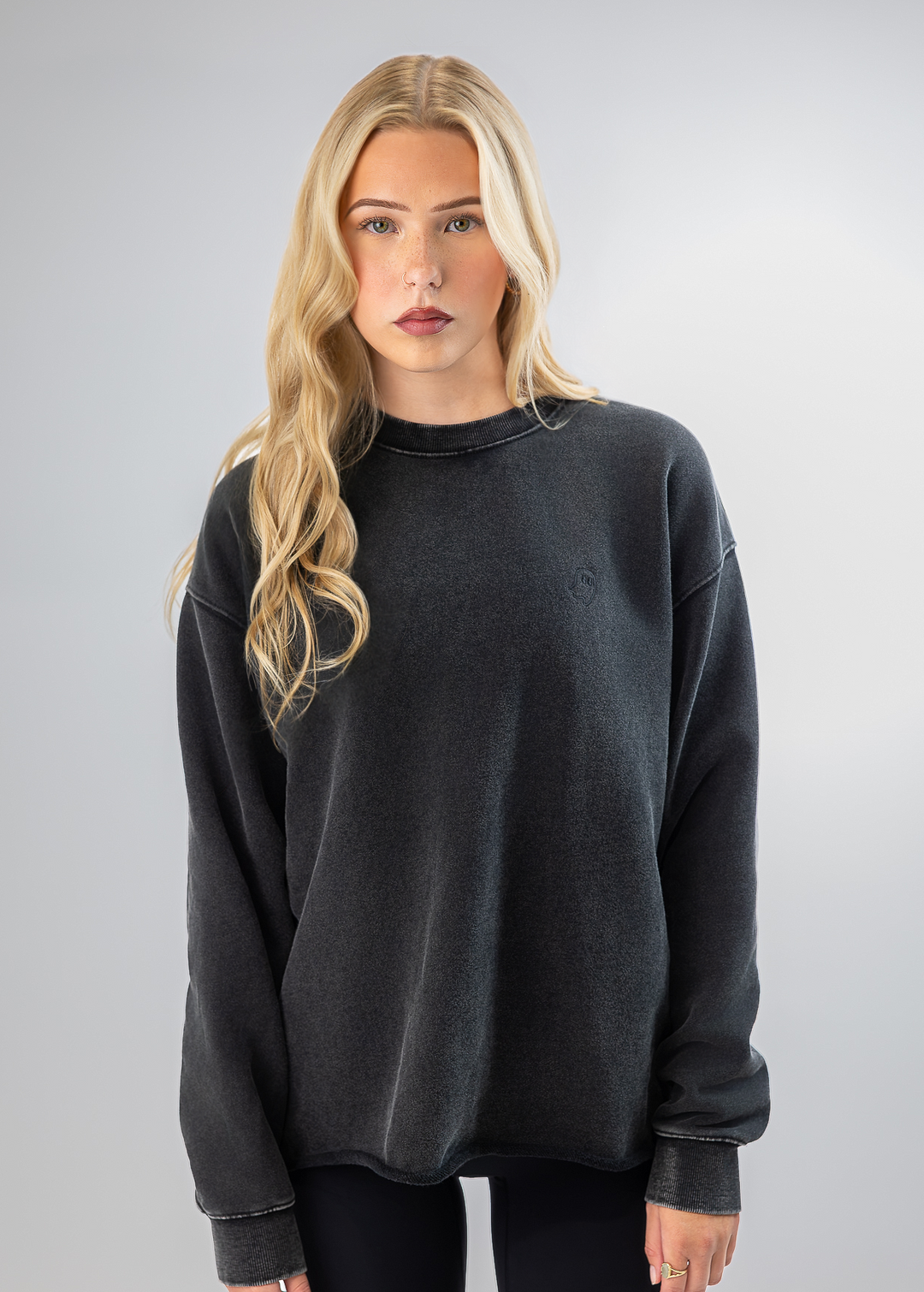 A blonde woman modeling a dark gray washed crewneck sweatshirt against a light gray studio background.