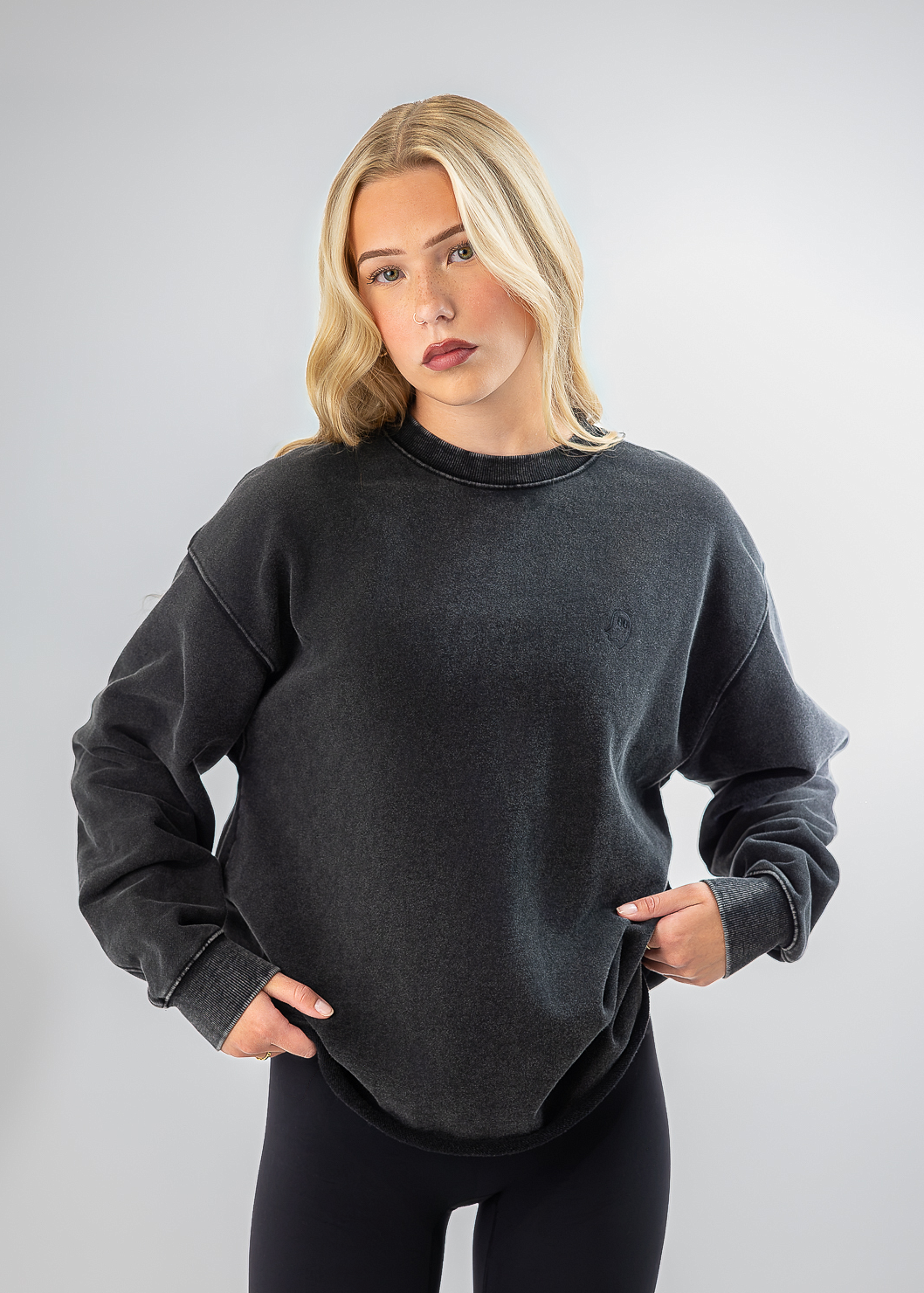 A blonde woman modeling a dark gray washed crewneck sweatshirt with black leggings against a light gray studio background.
