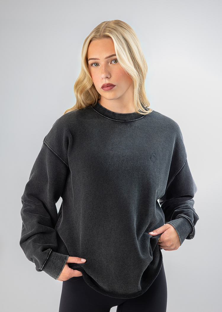 A blonde woman wearing a dark charcoal gray washed crewneck sweatshirt and black leggings, standing against a light gray studio background.