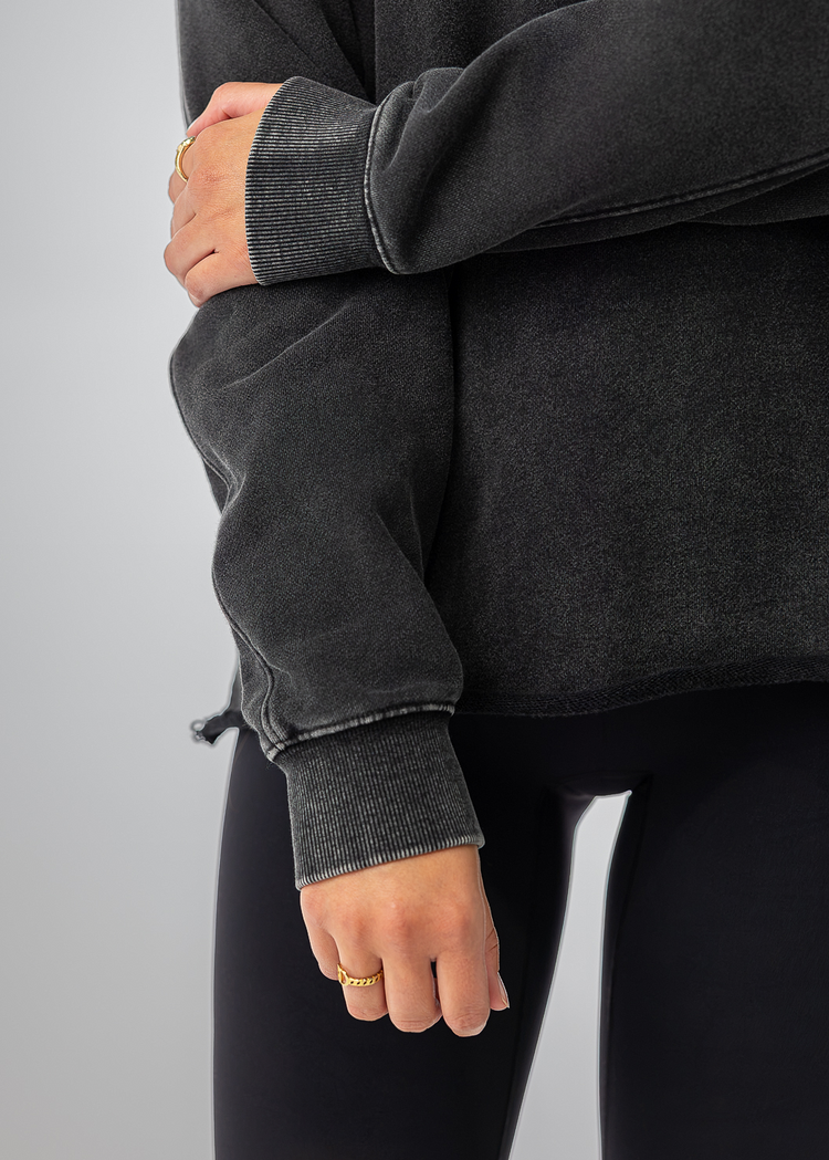 A close-up of a person wearing a dark charcoal gray oversized sweatshirt and black leggings, with one arm crossed over the other against a plain light gray studio background.