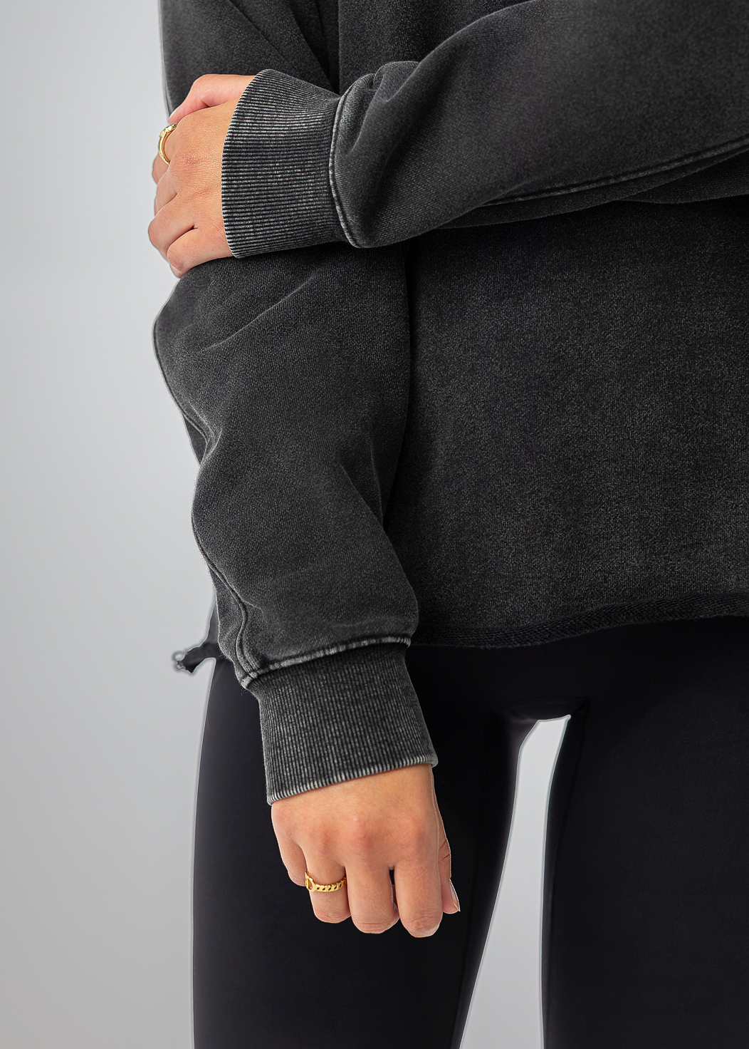 A close-up of a person wearing a dark charcoal gray oversized sweatshirt and black leggings, with one arm crossed over the other against a plain light gray studio background.