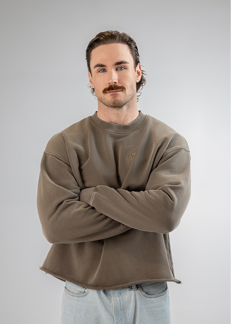 A man with a mustache wearing a brownish-gray crewneck sweatshirt and light blue jeans, standing with arms crossed against a light gray background.