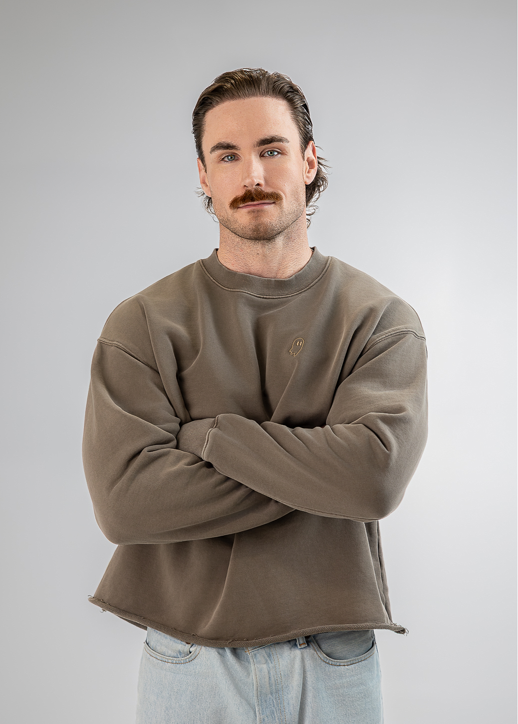 A man with a mustache wearing a brownish-gray crewneck sweatshirt and light blue jeans, standing with arms crossed against a light gray background.