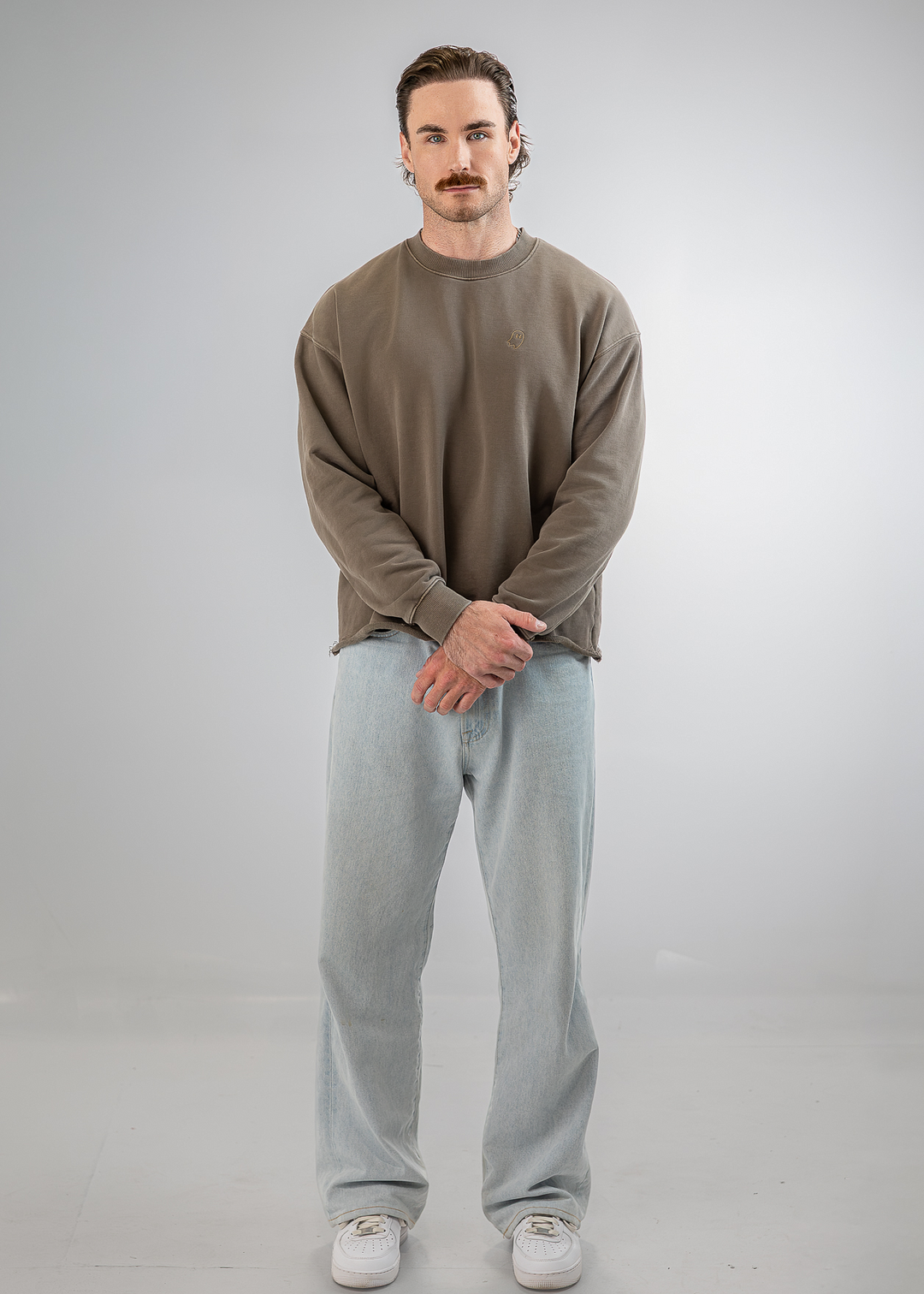 A man stands against a light gray studio background wearing a muted taupe-brown long-sleeve crewneck sweatshirt, light-wash blue jeans, and white sneakers.