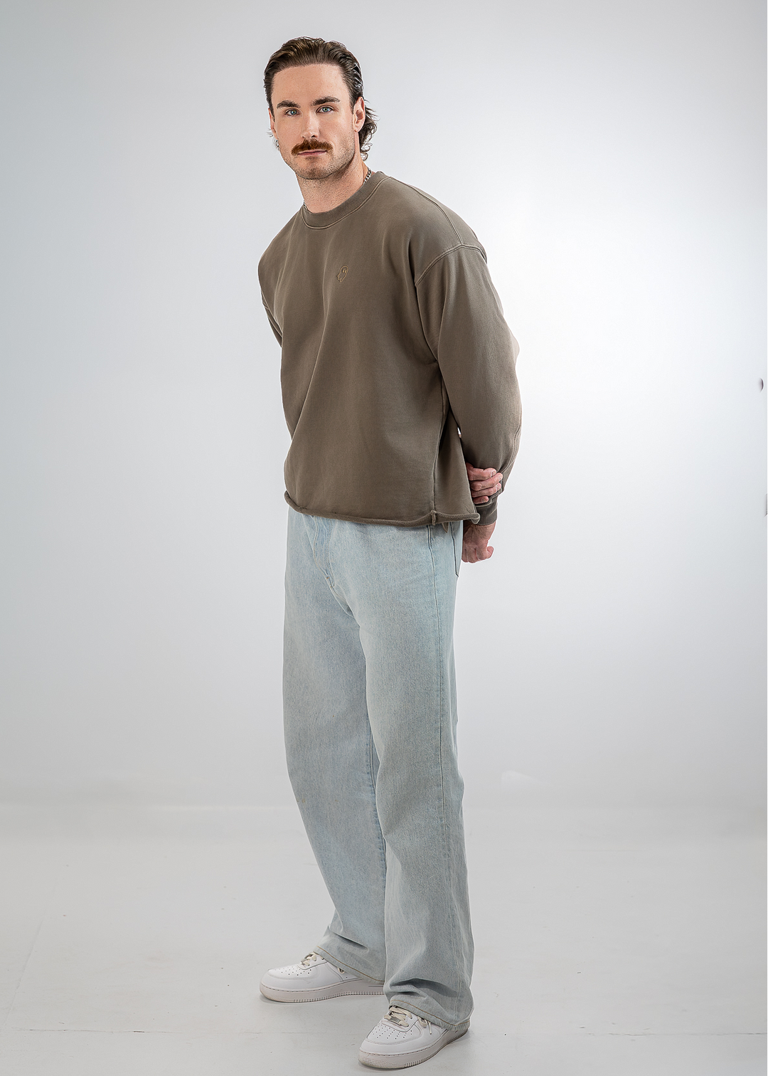 A man modeling a brown crewneck sweatshirt, light wash jeans, and white sneakers against a light gray studio background.