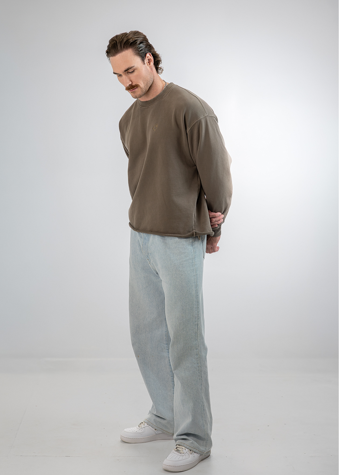 A man wearing a brown crewneck sweatshirt, light blue baggy jeans, and white sneakers, standing in a studio setting against a light gray background.