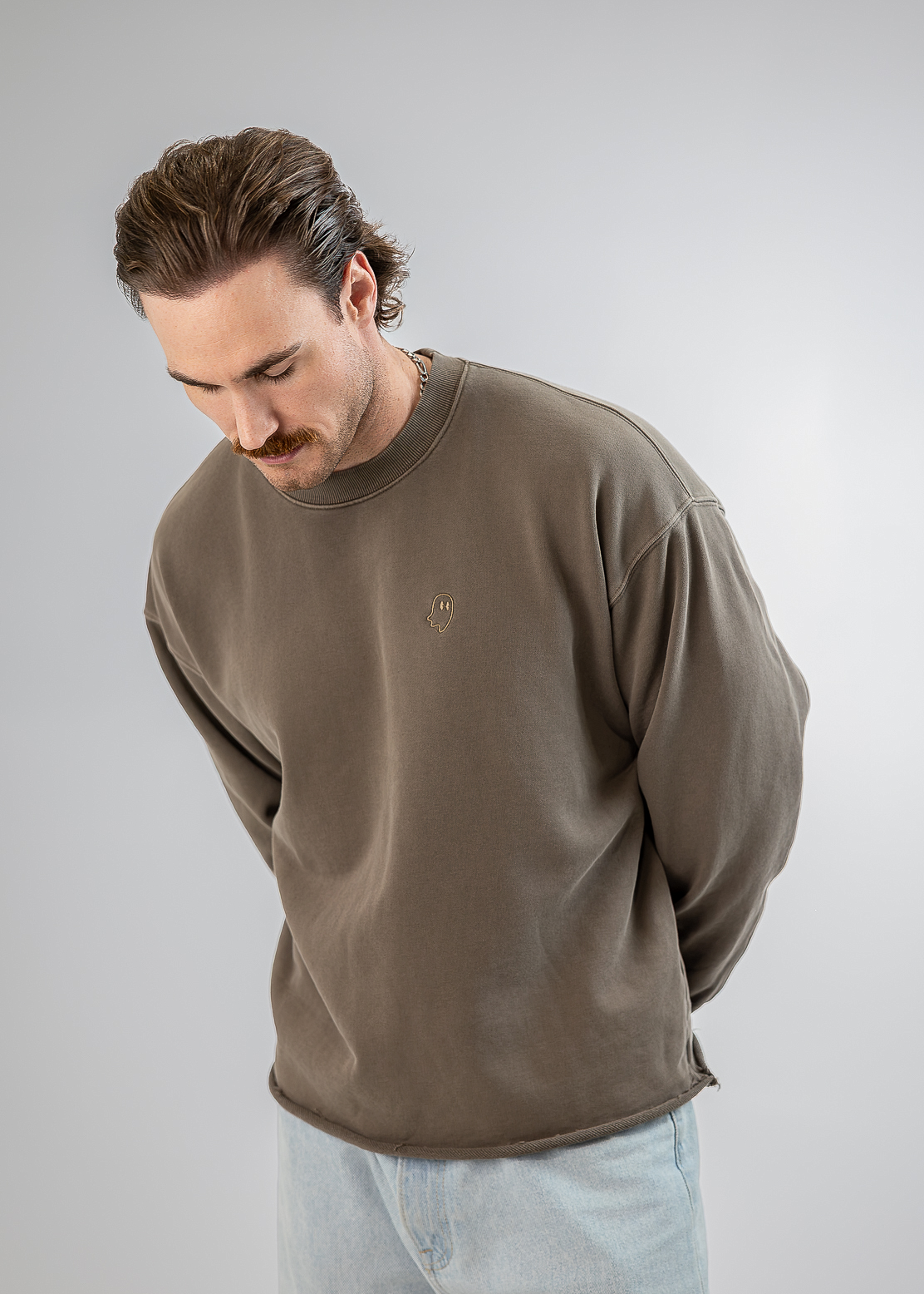 A man wearing a brown crewneck sweatshirt and light blue jeans, looking down against a neutral gray studio background.
