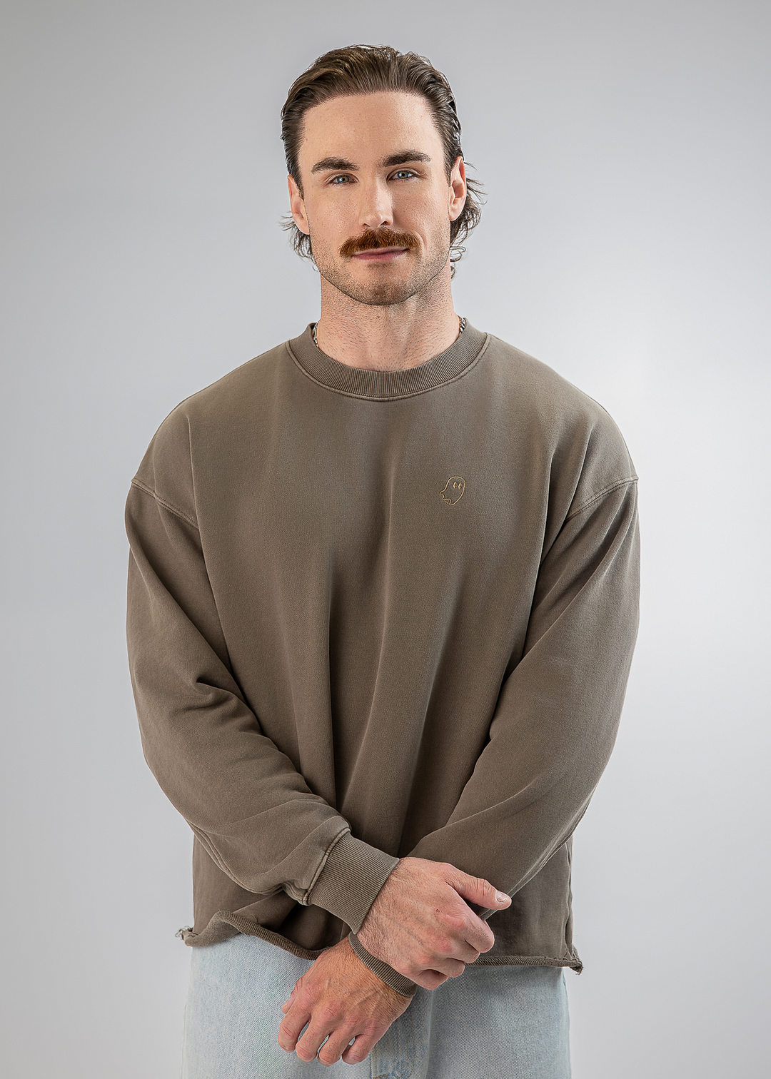 A man with a mustache wearing a taupe-brown crewneck sweatshirt and light blue jeans, standing against a neutral gray studio background.