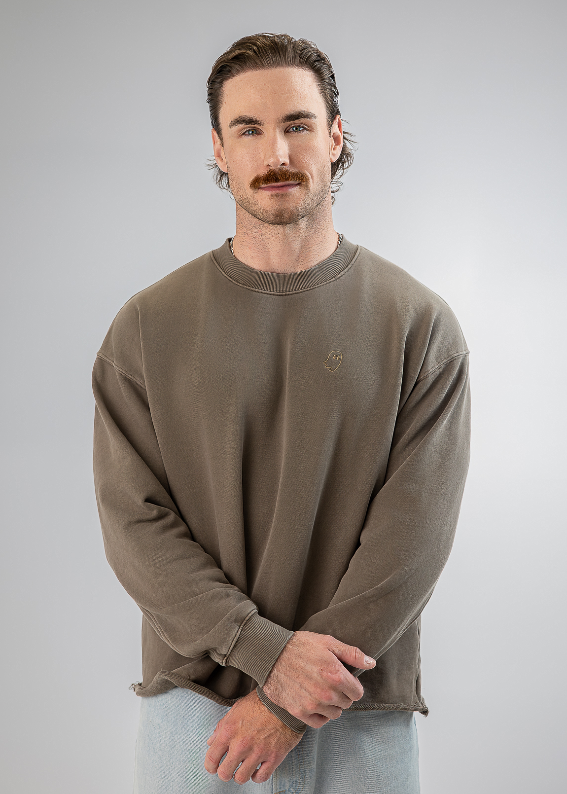 A man with a mustache wearing a taupe-brown crewneck sweatshirt and light blue jeans, standing against a neutral gray studio background.