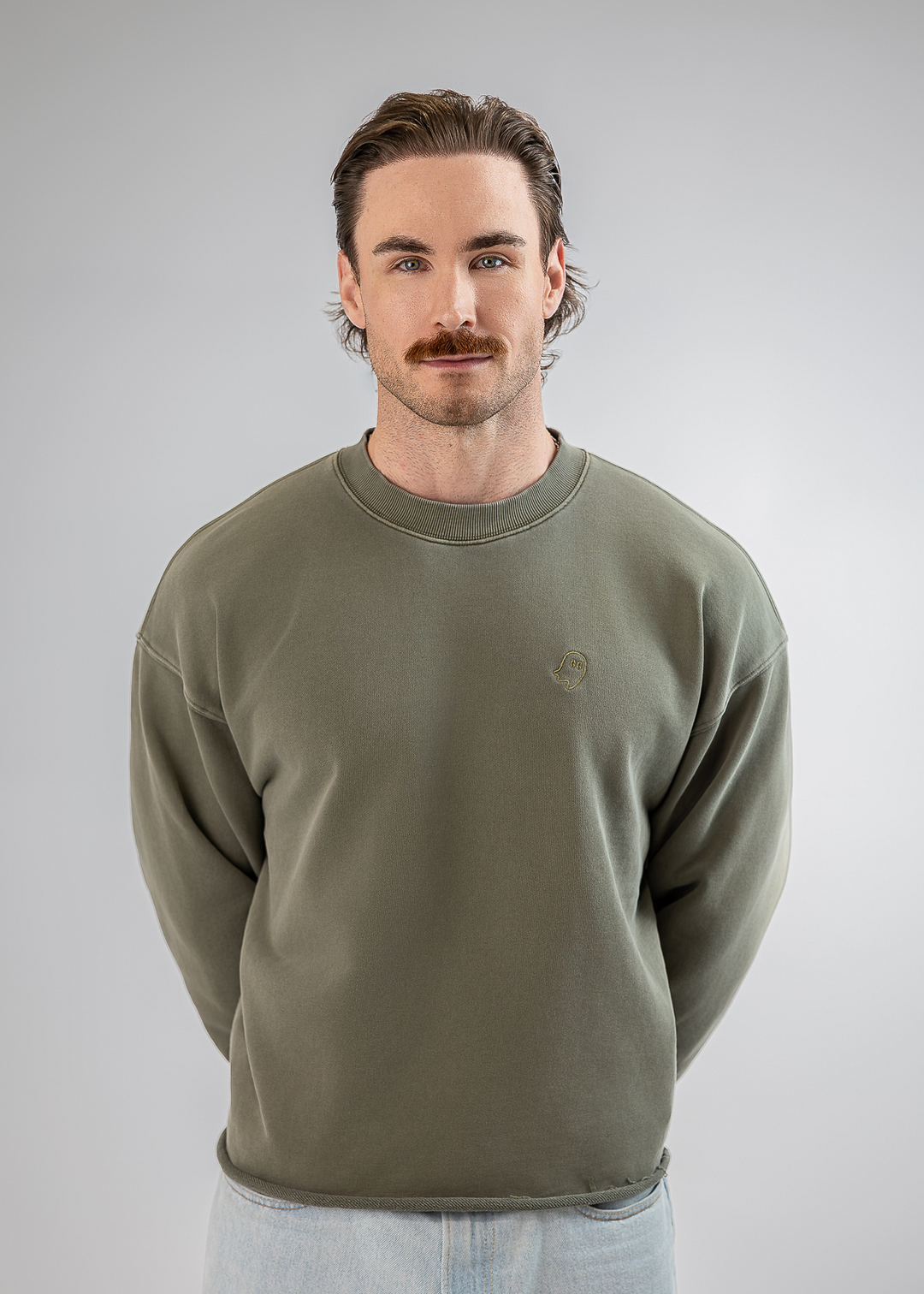 A man with a mustache wearing an olive green crewneck sweatshirt and light blue jeans, standing against a neutral gray studio background.