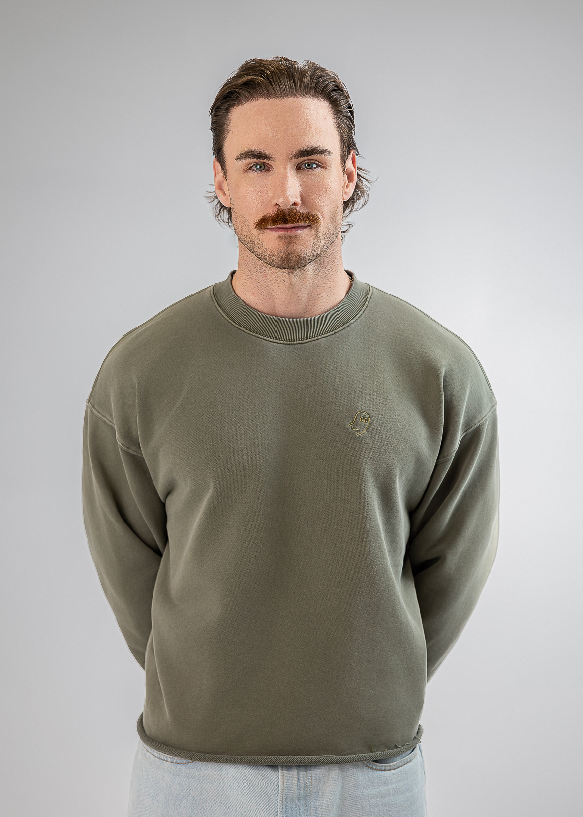 A man with a mustache wearing an olive green crewneck sweatshirt and light blue jeans, standing against a neutral gray studio background.