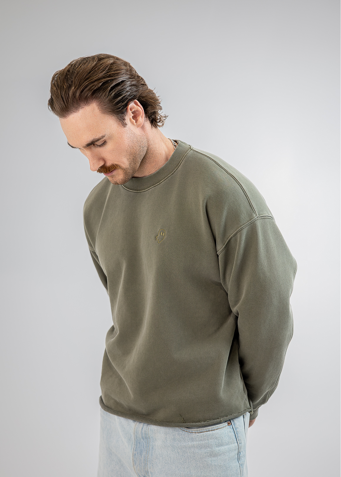 A man wearing an olive green crewneck sweatshirt and light blue jeans, looking down against a plain light gray studio background.