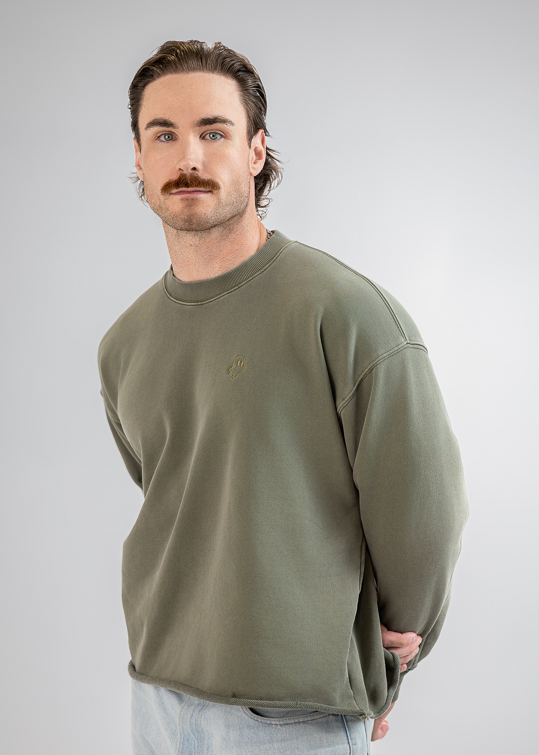 A man with a mustache wearing an olive green crewneck sweatshirt and light blue jeans, standing against a light gray studio background.