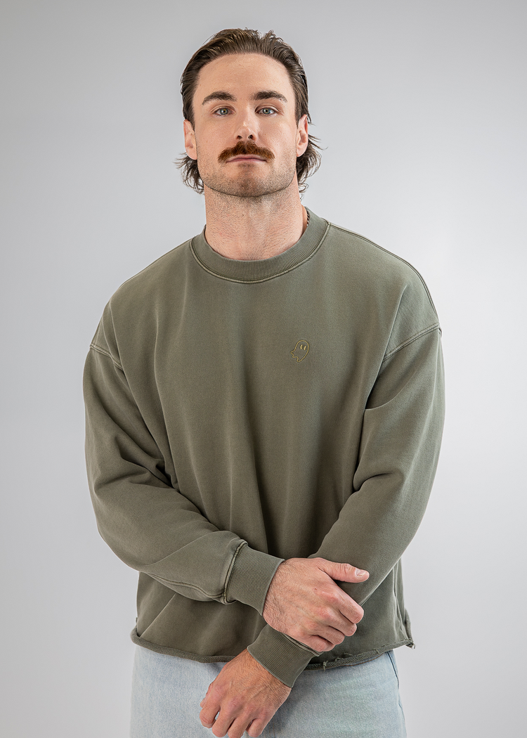 A man wearing an olive green crewneck sweatshirt and light blue jeans, standing against a neutral gray studio background.
