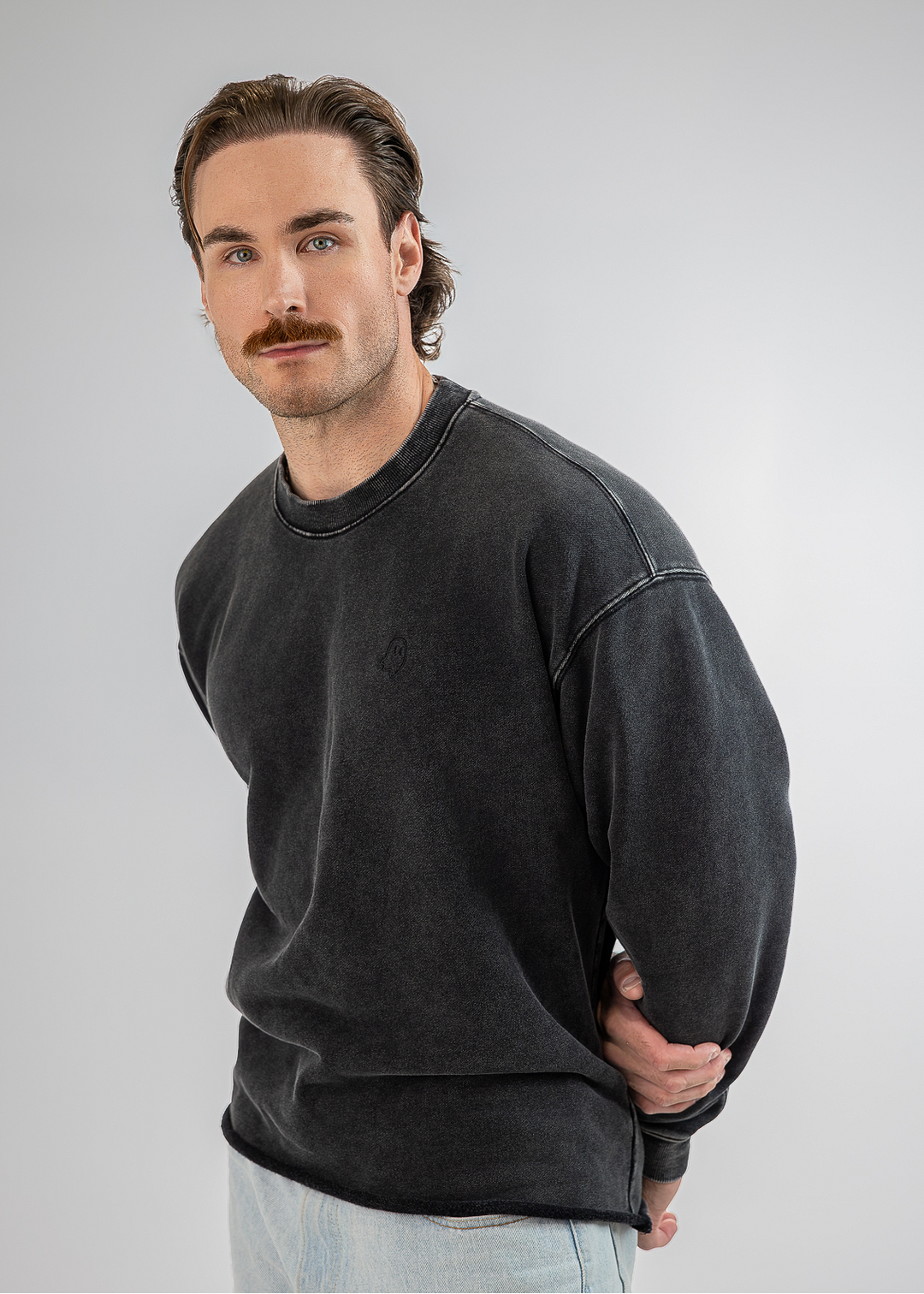 A man wearing a washed black crewneck sweatshirt and light blue jeans, standing against a neutral gray studio background.