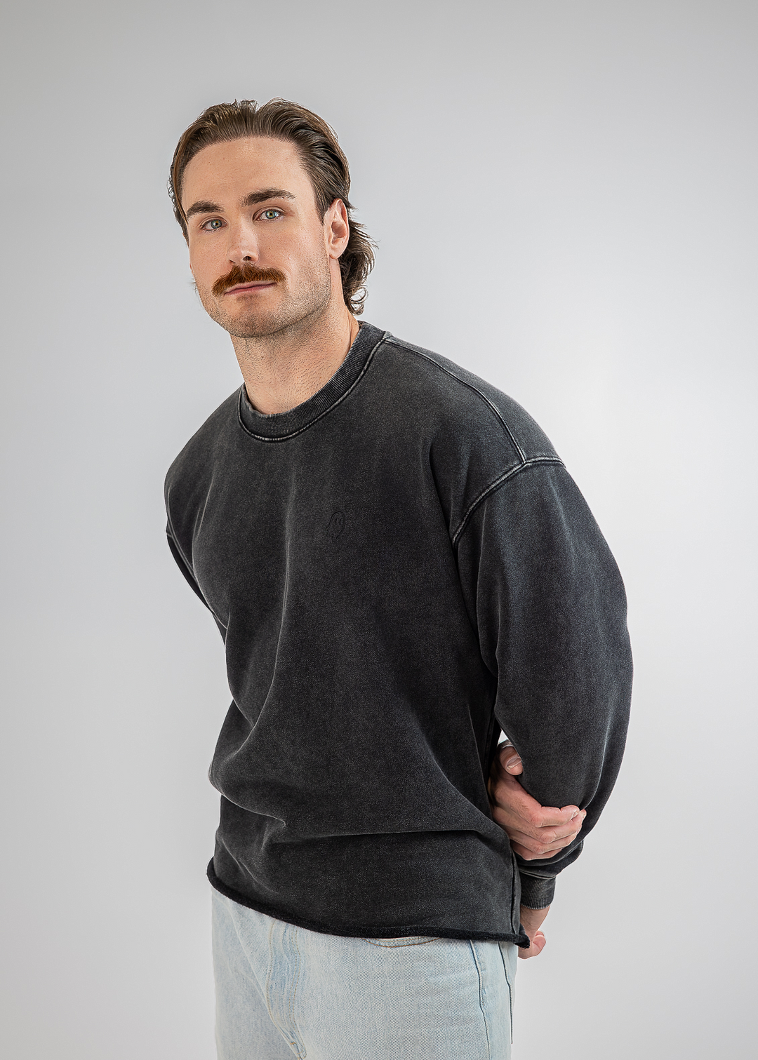 A man wearing a washed black crewneck sweatshirt and light blue jeans, standing against a neutral gray studio background.