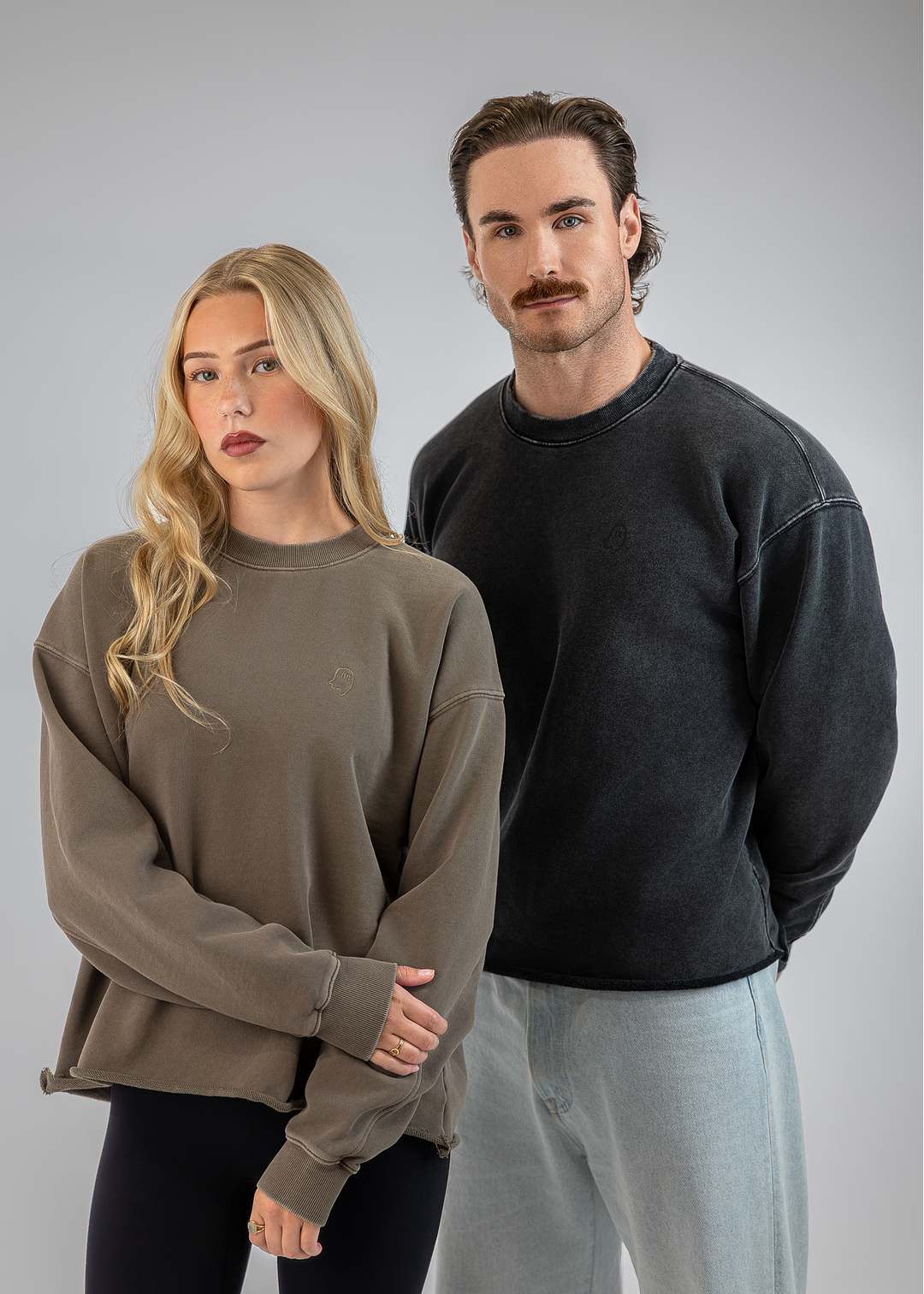 A man and a woman modeling oversized crewneck sweatshirts against a solid gray studio background. The woman wears a muted taupe-brown sweatshirt, and the man wears a dark charcoal gray sweatshirt.
