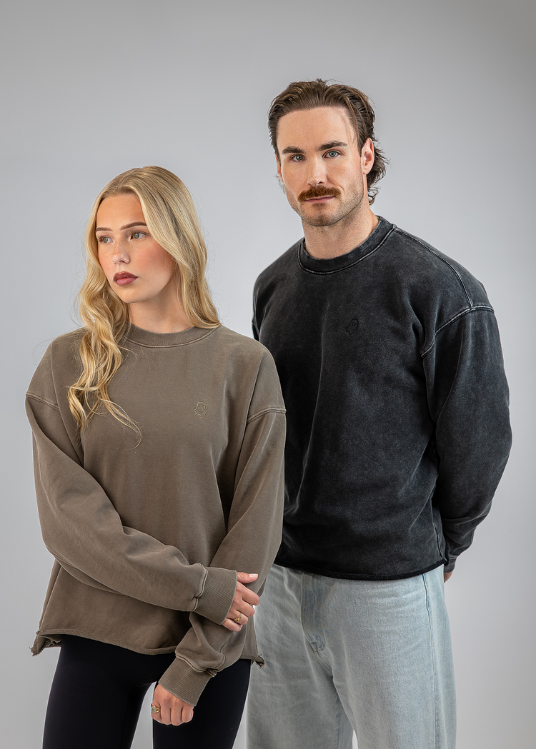 A man and a woman modeling oversized crewneck sweatshirts against a solid gray studio background. The woman on the left wears a muted taupe-brown sweatshirt, and the man on the right wears a washed charcoal-black sweatshirt.