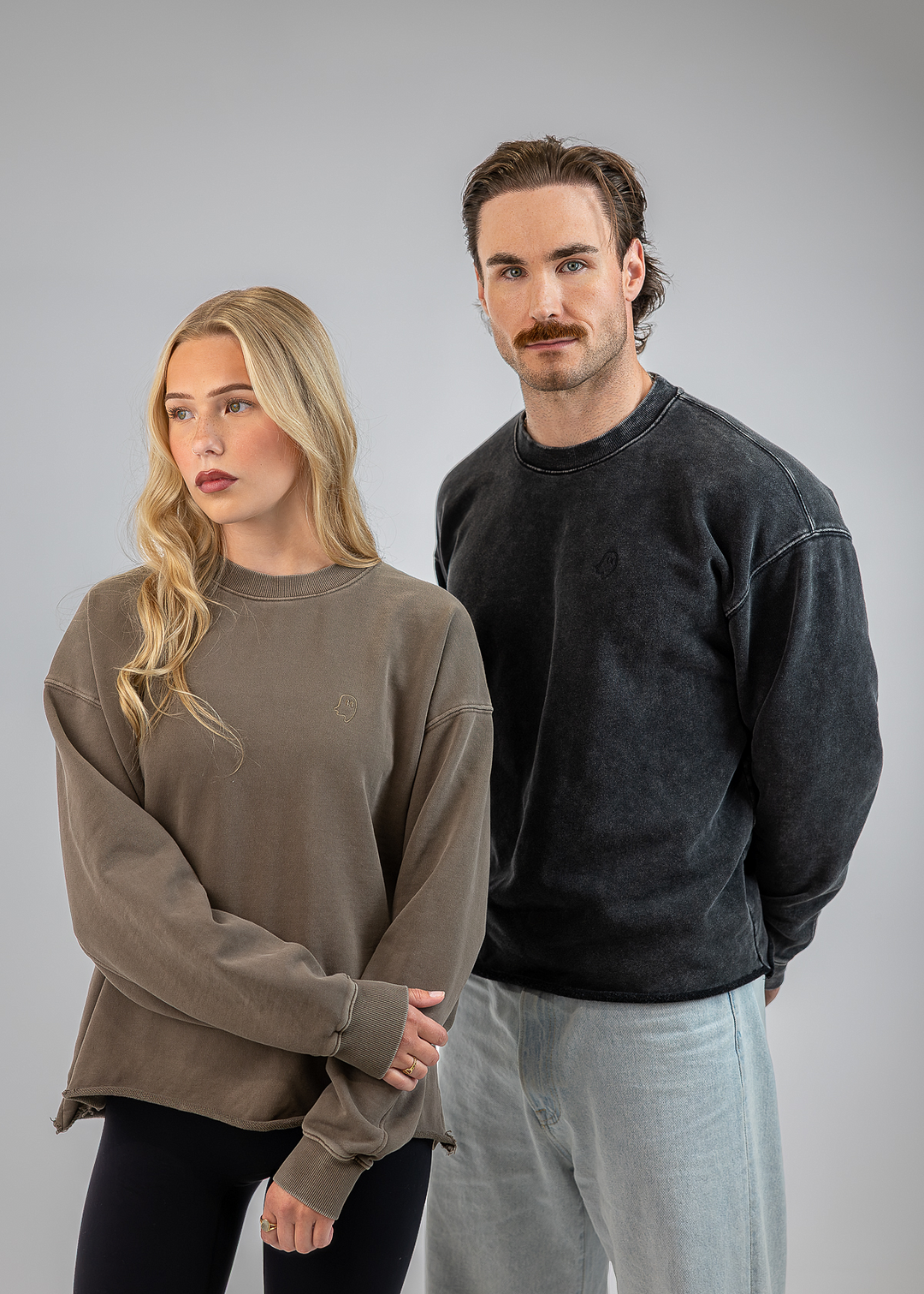 A man and a woman modeling oversized crewneck sweatshirts against a solid gray studio background. The woman on the left wears a muted taupe-brown sweatshirt, and the man on the right wears a washed charcoal-black sweatshirt.