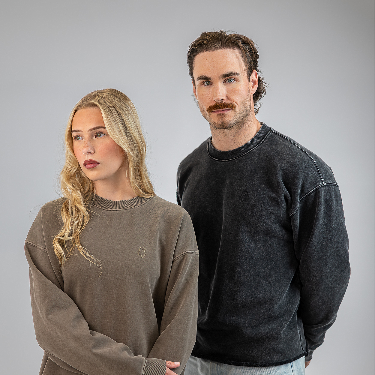 A man and a woman modeling oversized crewneck sweatshirts against a solid gray studio background. The woman on the left wears a muted taupe-brown sweatshirt, and the man on the right wears a washed charcoal-black sweatshirt.