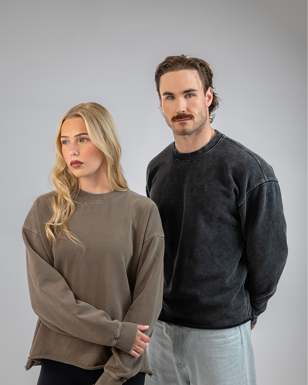 A man and a woman modeling oversized crewneck sweatshirts against a solid gray studio background. The woman on the left wears a muted taupe-brown sweatshirt, and the man on the right wears a washed charcoal-black sweatshirt.