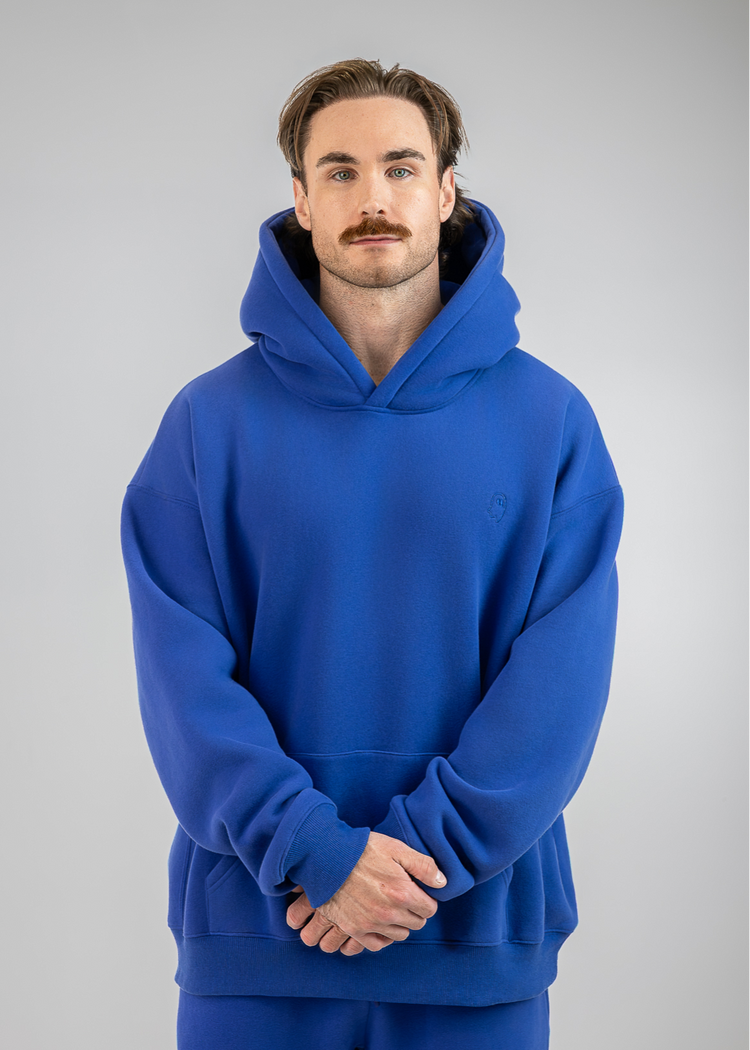 A man wearing a royal blue pullover hoodie and matching sweatpants, standing against a neutral gray studio background.