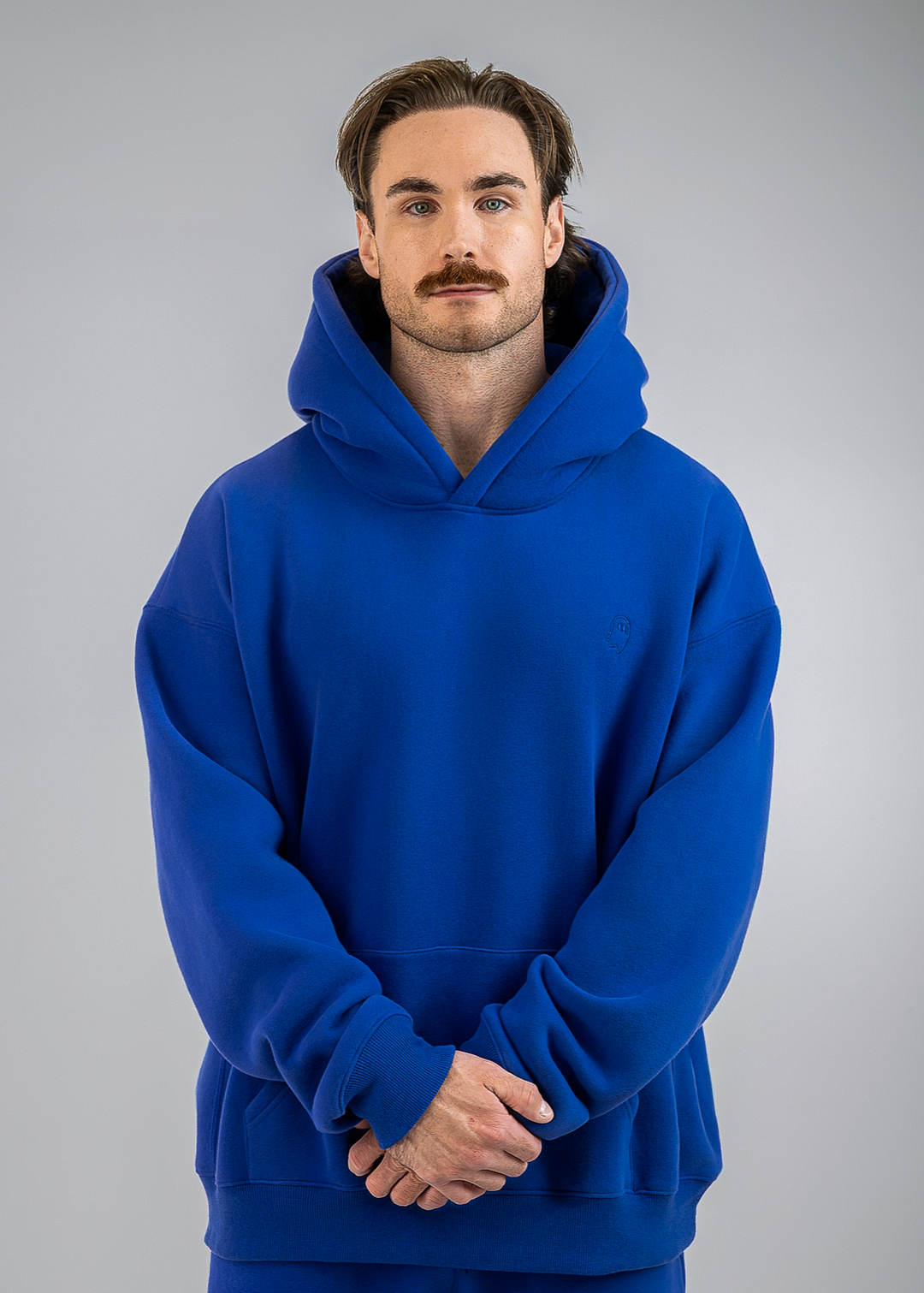A man wearing a bright royal blue pullover hoodie and matching blue pants, standing against a neutral gray studio background.