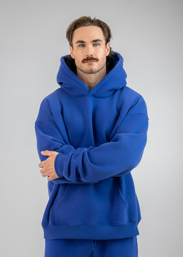 A man with a mustache wearing a royal blue pullover hoodie and matching blue sweatpants, standing against a neutral gray studio background with his arms crossed.
