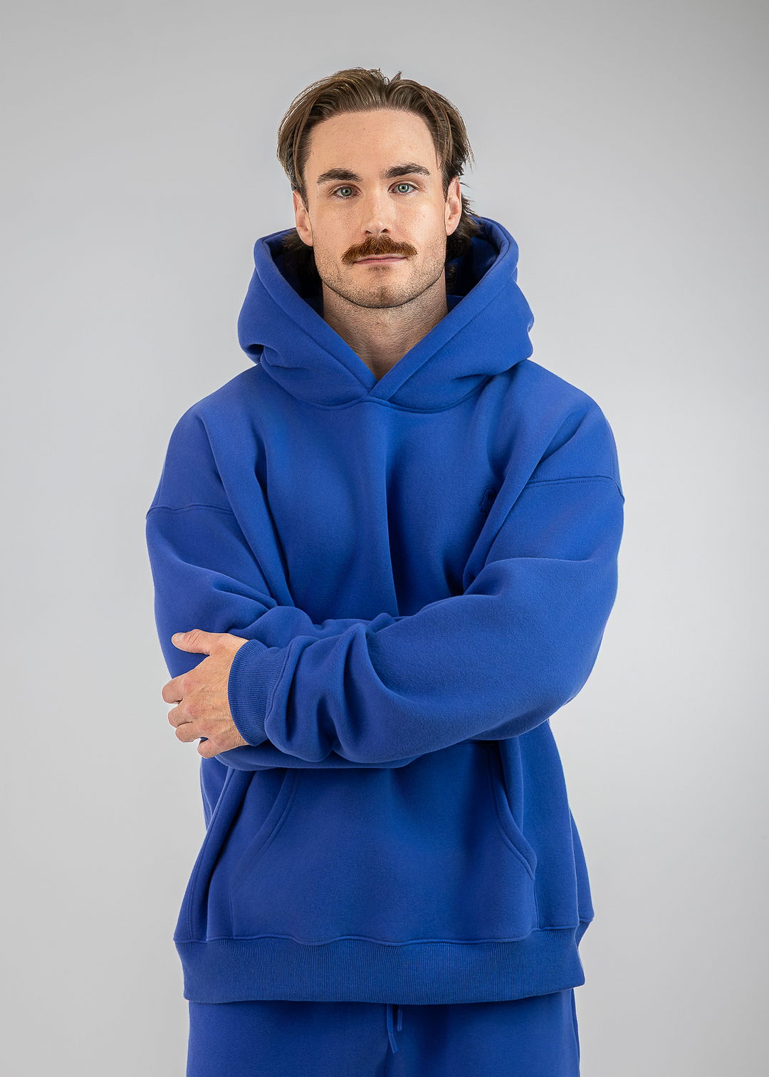 A man with a mustache wearing a royal blue pullover hoodie and matching blue sweatpants, standing against a neutral gray studio background with his arms crossed.