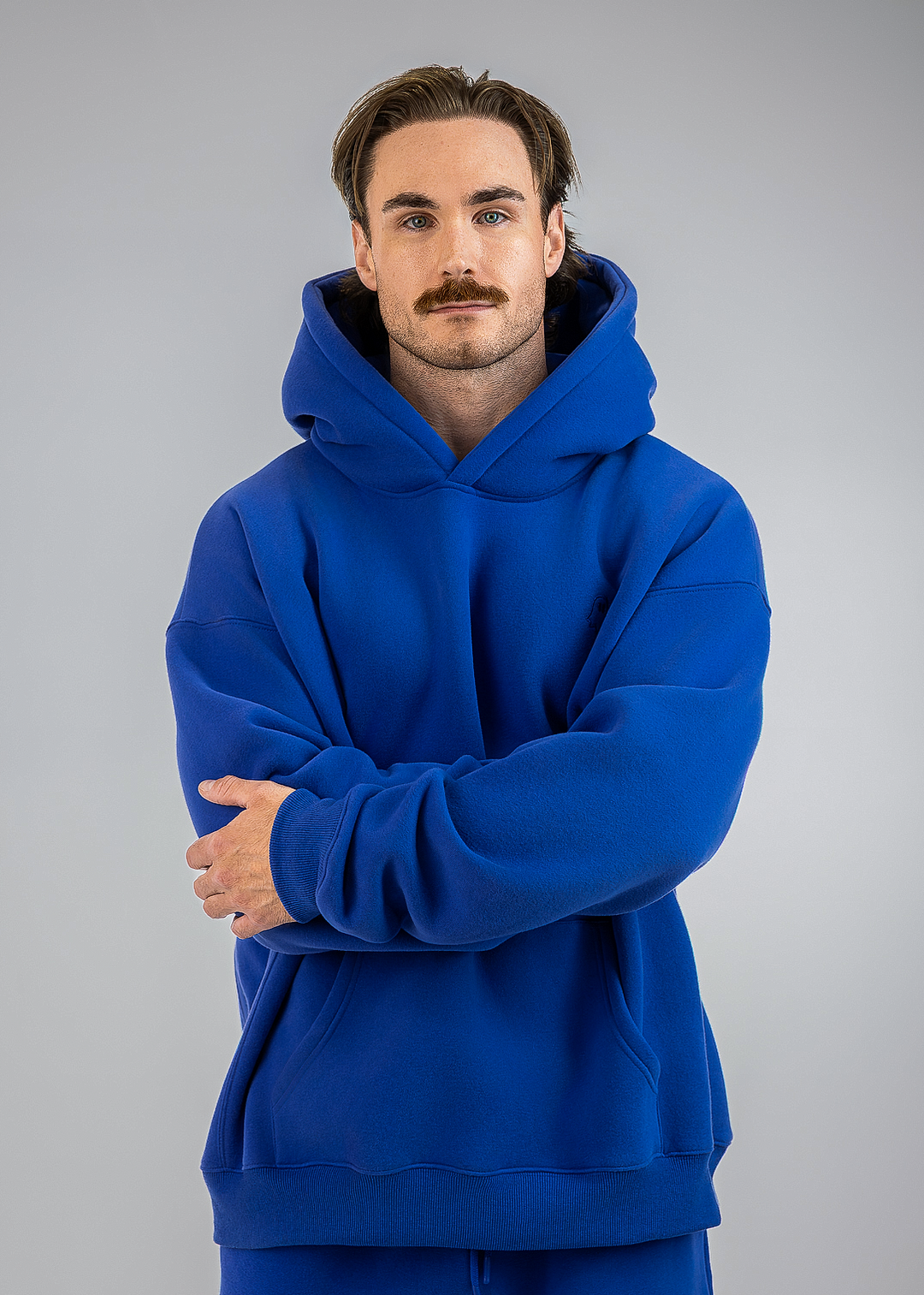 A man with a mustache wearing a vibrant royal blue hoodie and matching sweatpants, standing against a solid gray studio background with his arms crossed.