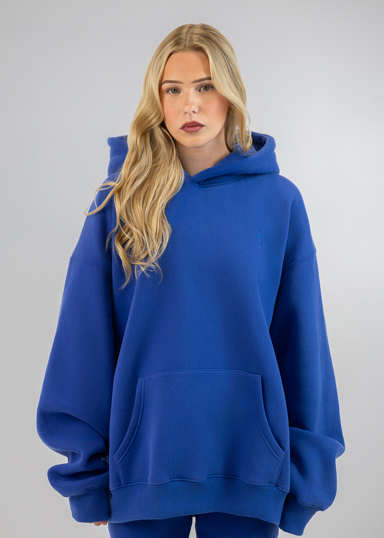 A blonde woman wearing an oversized royal blue pullover hoodie with a front kangaroo pocket, paired with matching blue leggings, set against a plain light gray studio background.