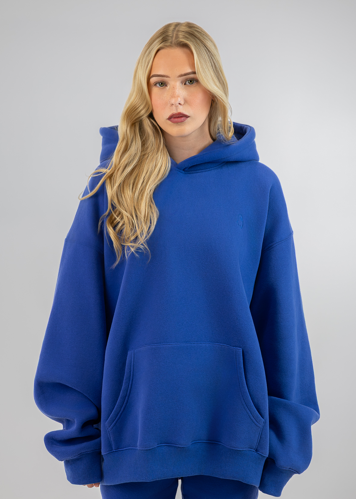 A blonde woman wearing an oversized royal blue pullover hoodie with a front kangaroo pocket, paired with matching blue leggings, set against a plain light gray studio background.
