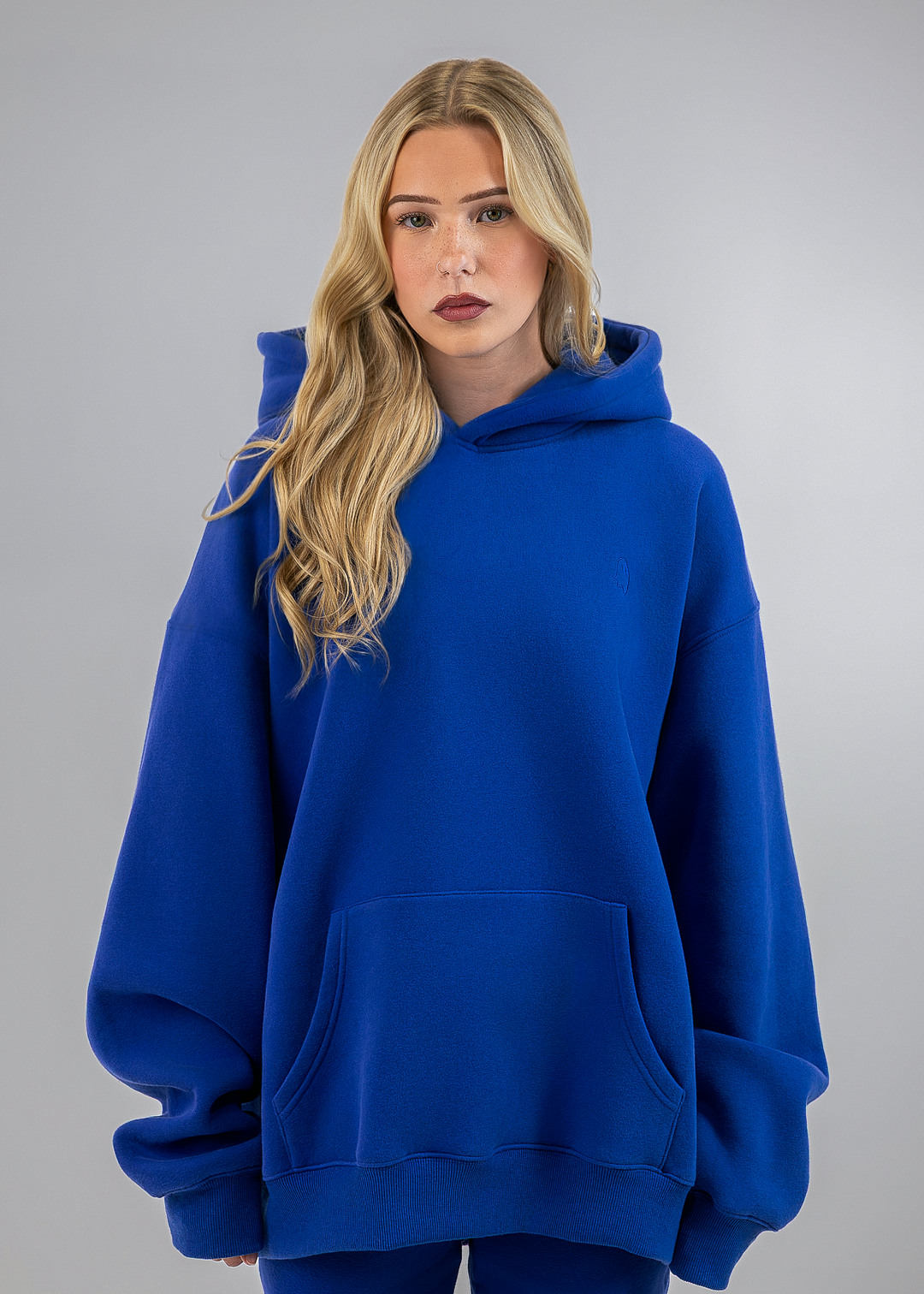 A blonde woman wearing an oversized royal blue hoodie with a front kangaroo pocket, photographed against a neutral gray studio background.