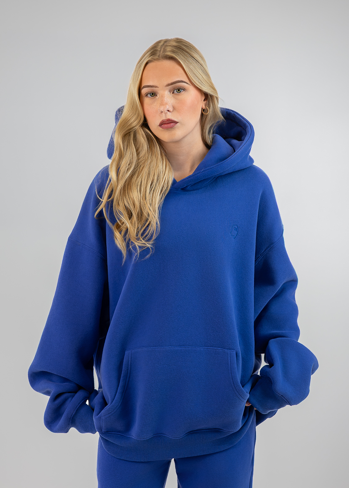 A blonde woman wearing an oversized royal blue hoodie and matching blue sweatpants, standing against a light gray studio background.