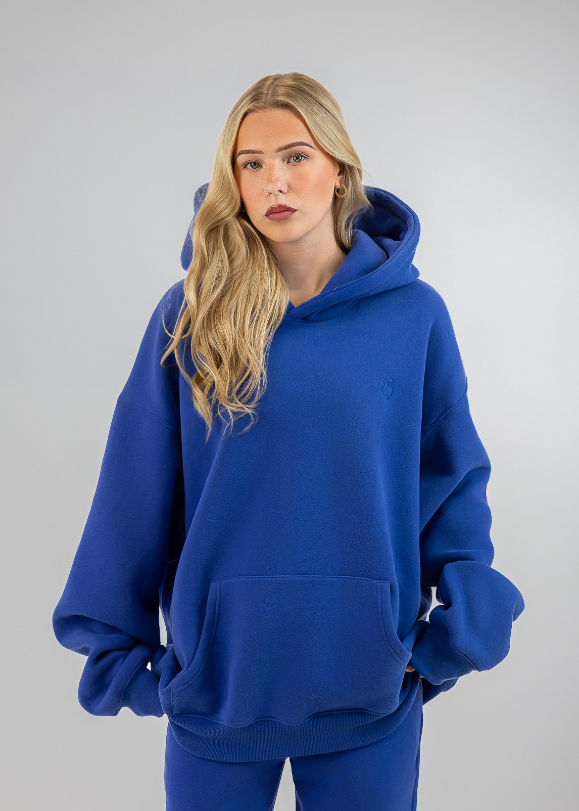 A blonde woman wearing an oversized royal blue hoodie and matching blue sweatpants, standing against a light gray studio background.