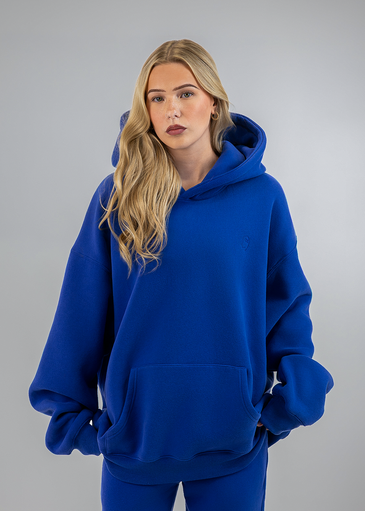 A blonde woman wearing an oversized royal blue hoodie and matching blue sweatpants, standing against a neutral gray studio background.