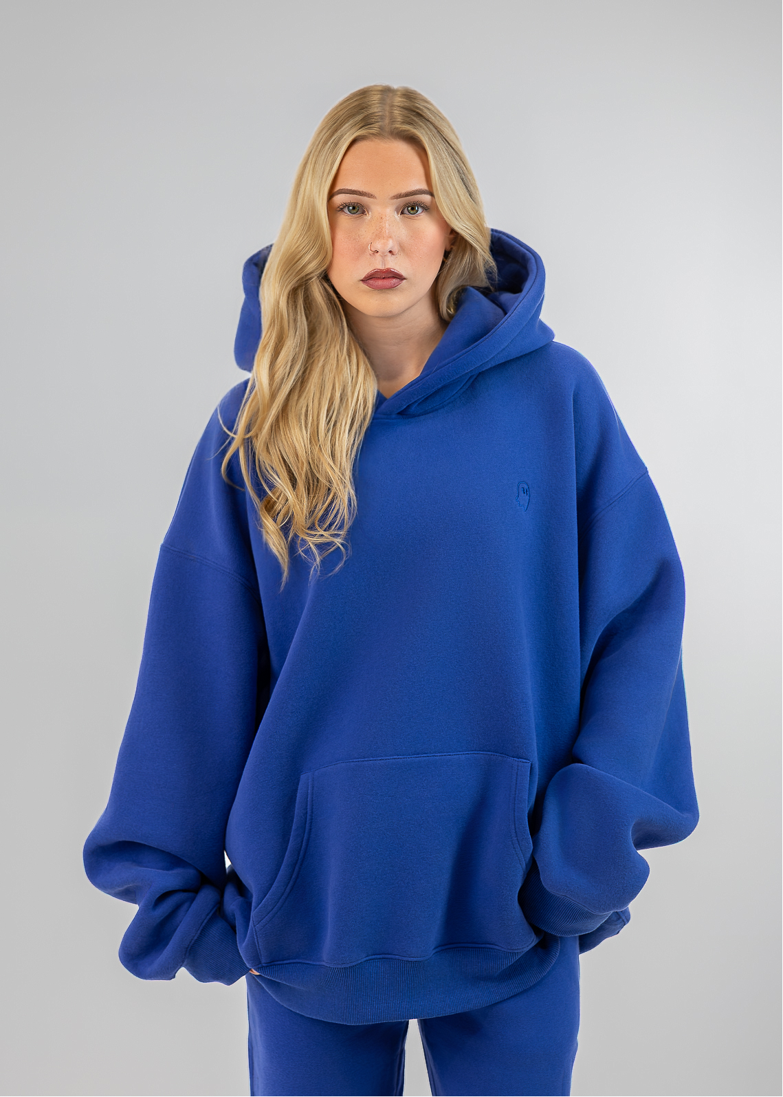 A blonde woman wearing an oversized royal blue hoodie and matching blue sweatpants, standing against a neutral light gray studio background.