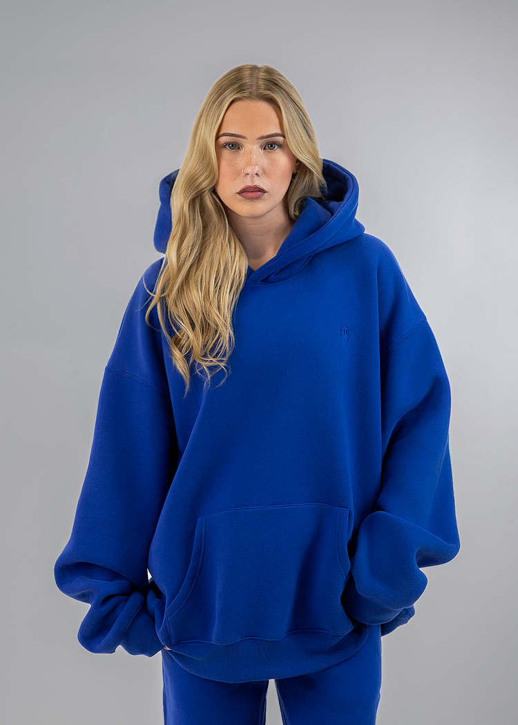 A blonde woman modeling an oversized royal blue hoodie and matching blue sweatpants against a solid gray studio background.