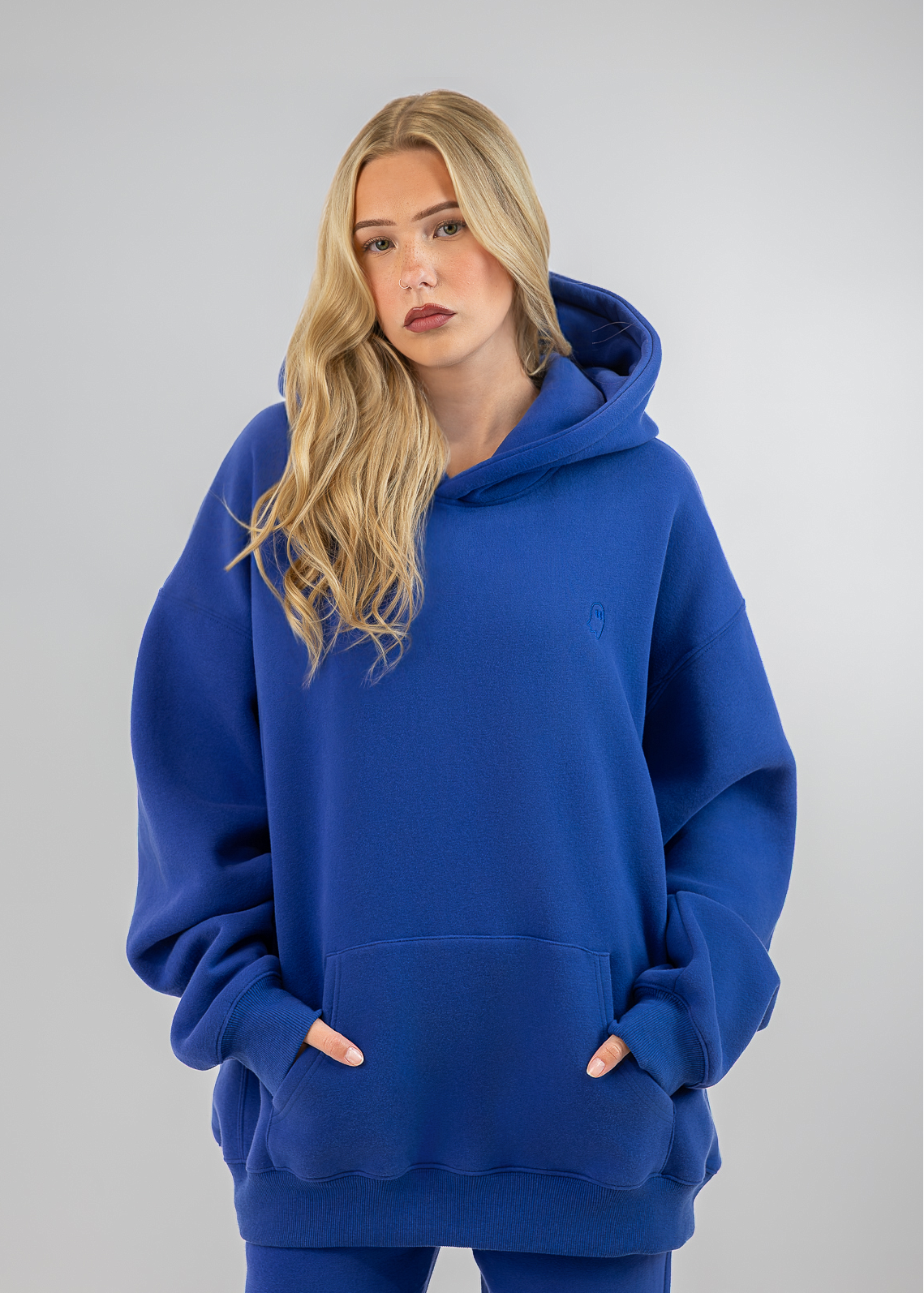 A blonde woman wearing an oversized royal blue hoodie and matching blue pants, standing against a neutral gray studio background.