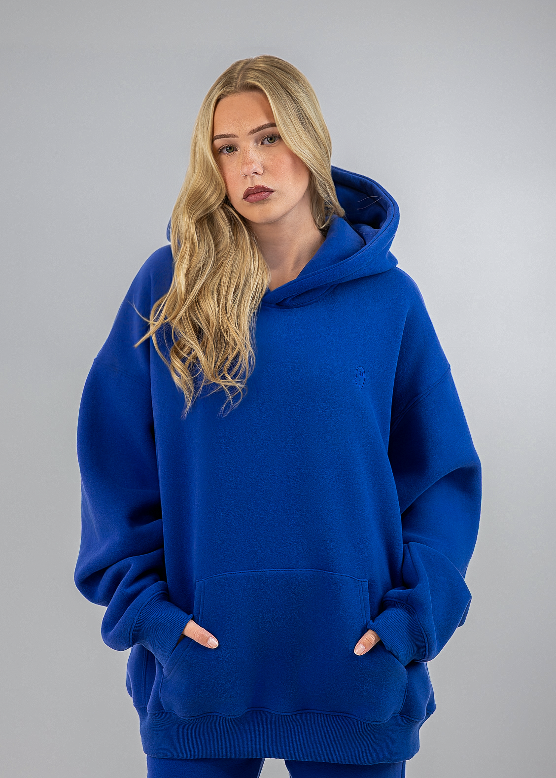 A blonde woman wearing an oversized royal blue hoodie and matching blue pants, standing against a neutral gray studio background.