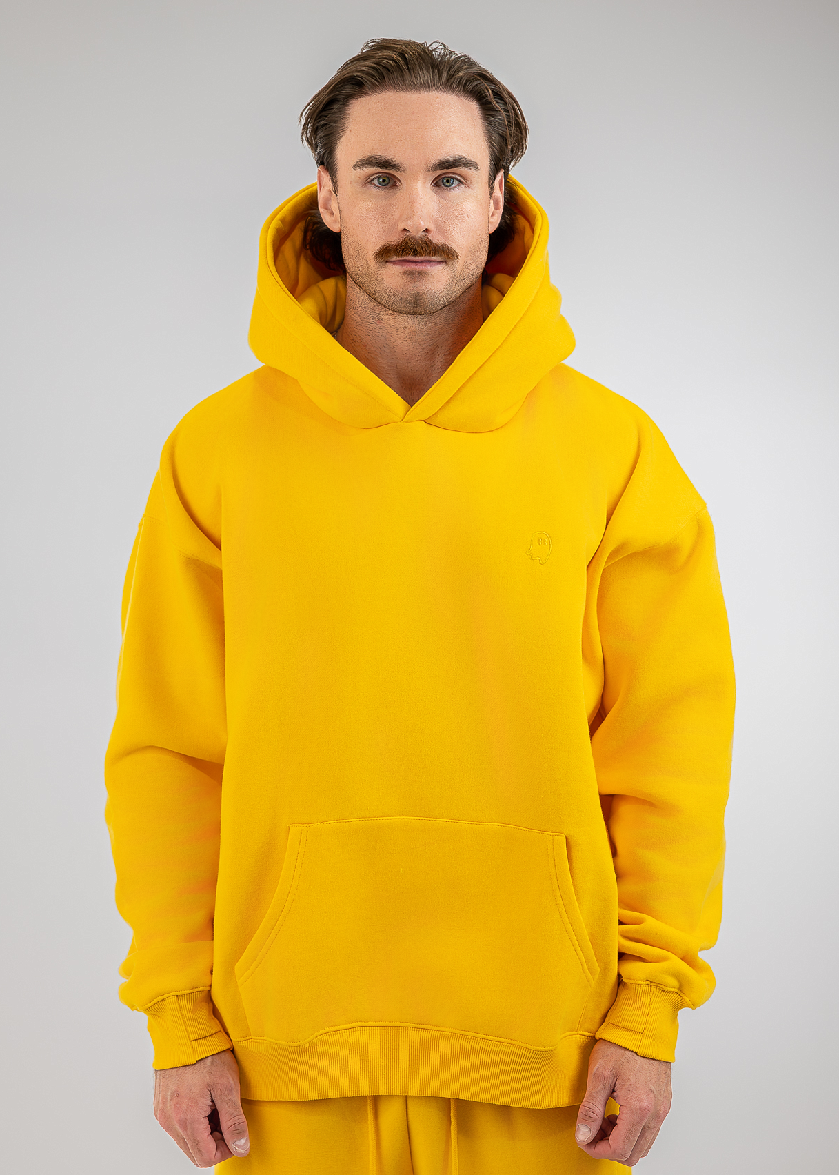 A man wearing a bright yellow pullover hoodie and matching yellow sweatpants, standing against a neutral light gray studio background.