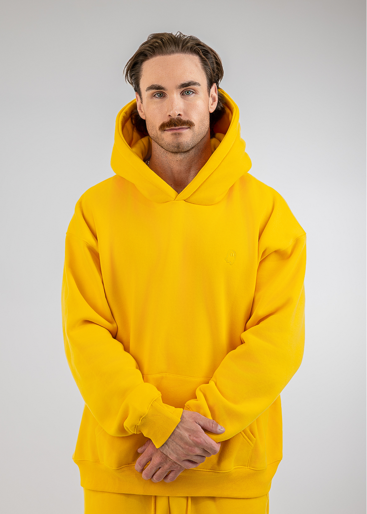 A man wearing a bright yellow pullover hoodie and matching sweatpants, standing against a neutral light gray studio background.
