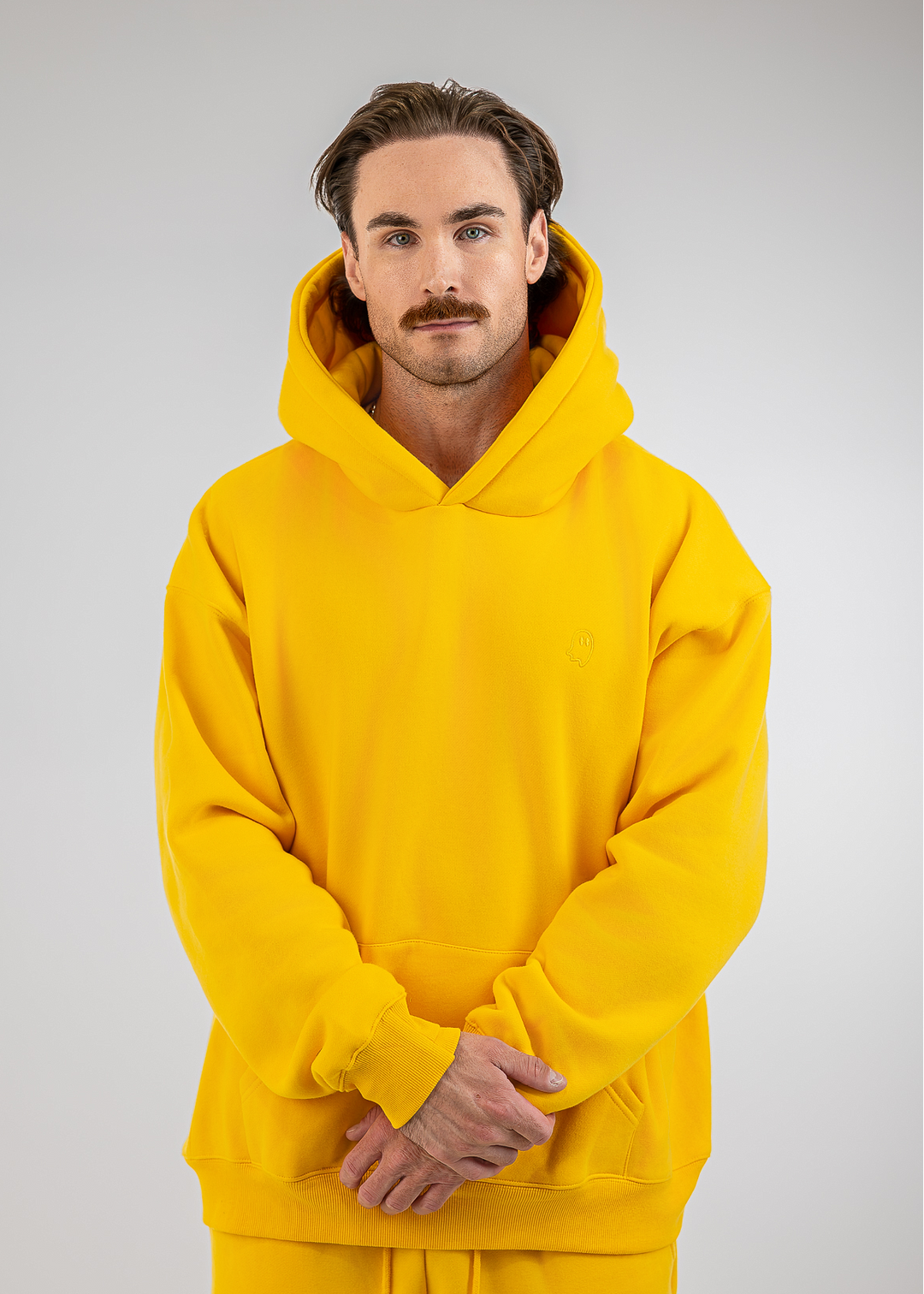 A man wearing a bright yellow pullover hoodie and matching sweatpants, standing against a neutral light gray studio background.