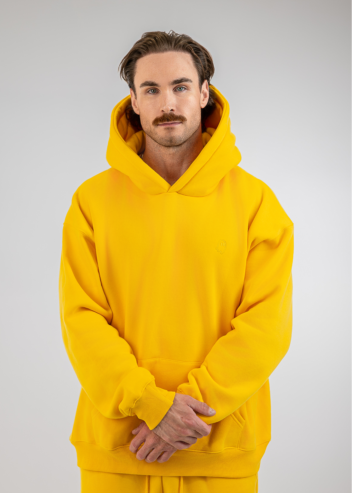 A man wearing a bright yellow pullover hoodie and matching sweatpants, standing against a neutral light gray studio background.